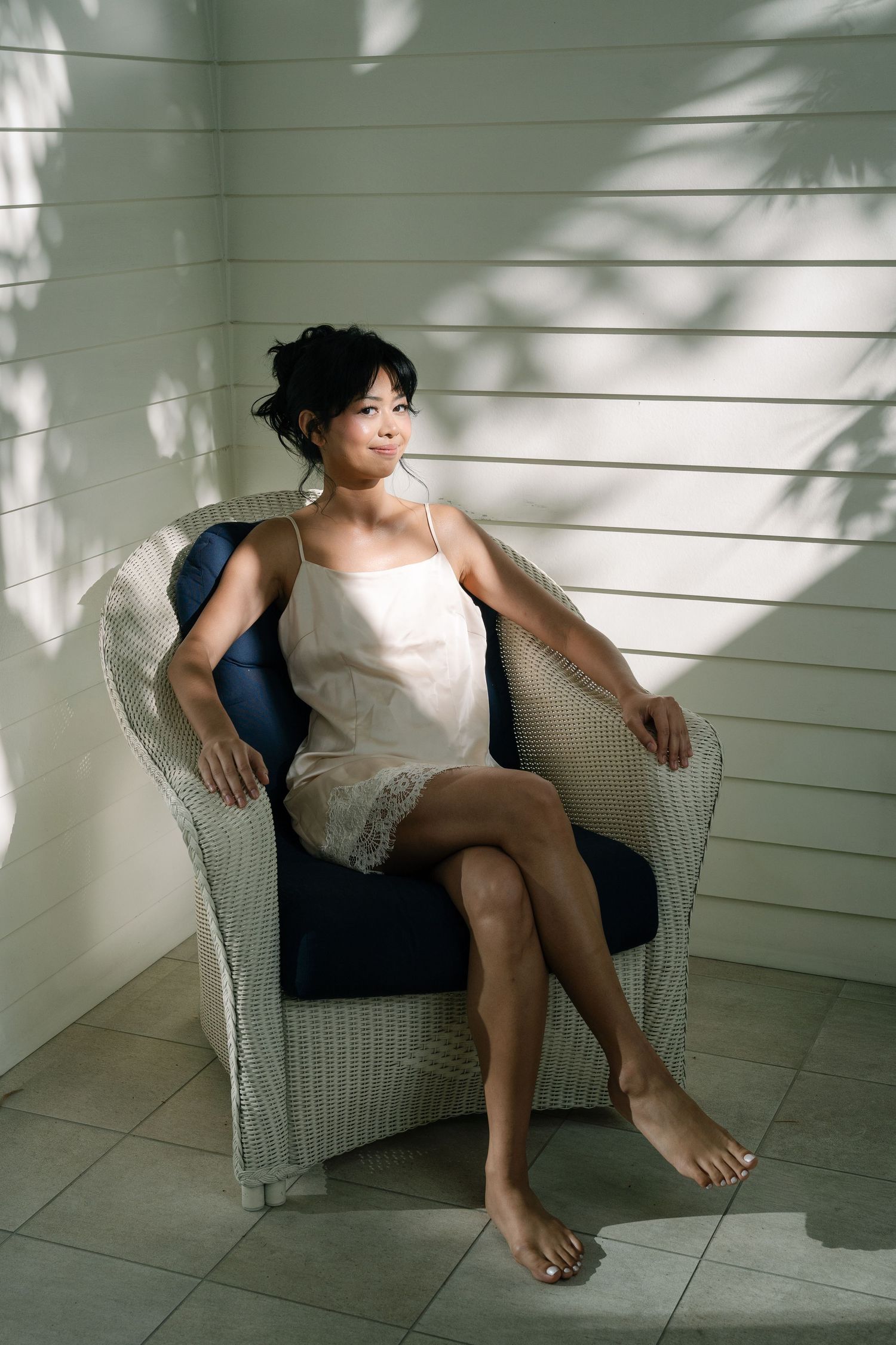 A young Asian woman in a white slip dress sits relaxed in a wicker chair, bathed in natural sunlight.