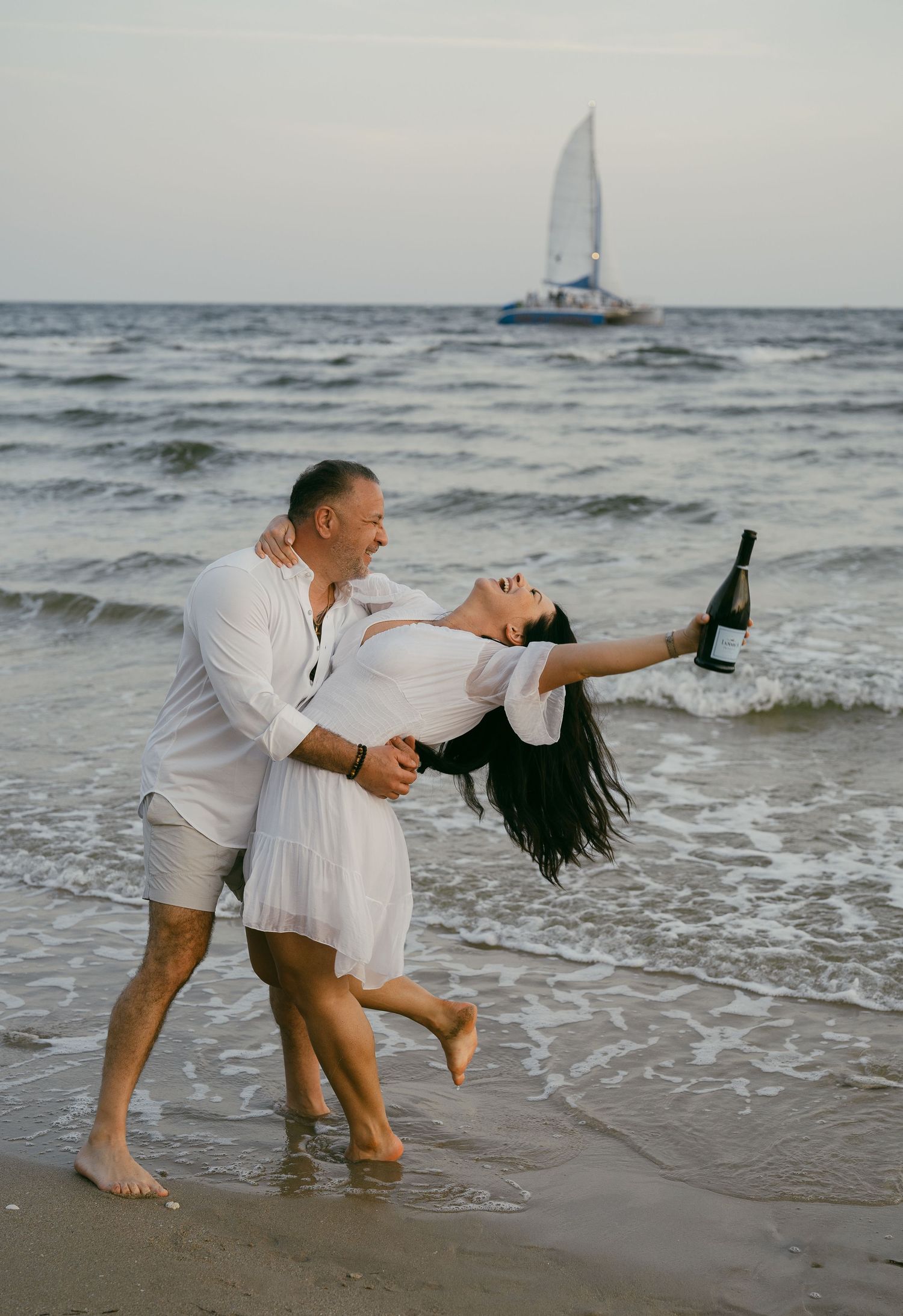 Couple playfully interacting on a beach at dusk with gentle waves and a boat in the background.
