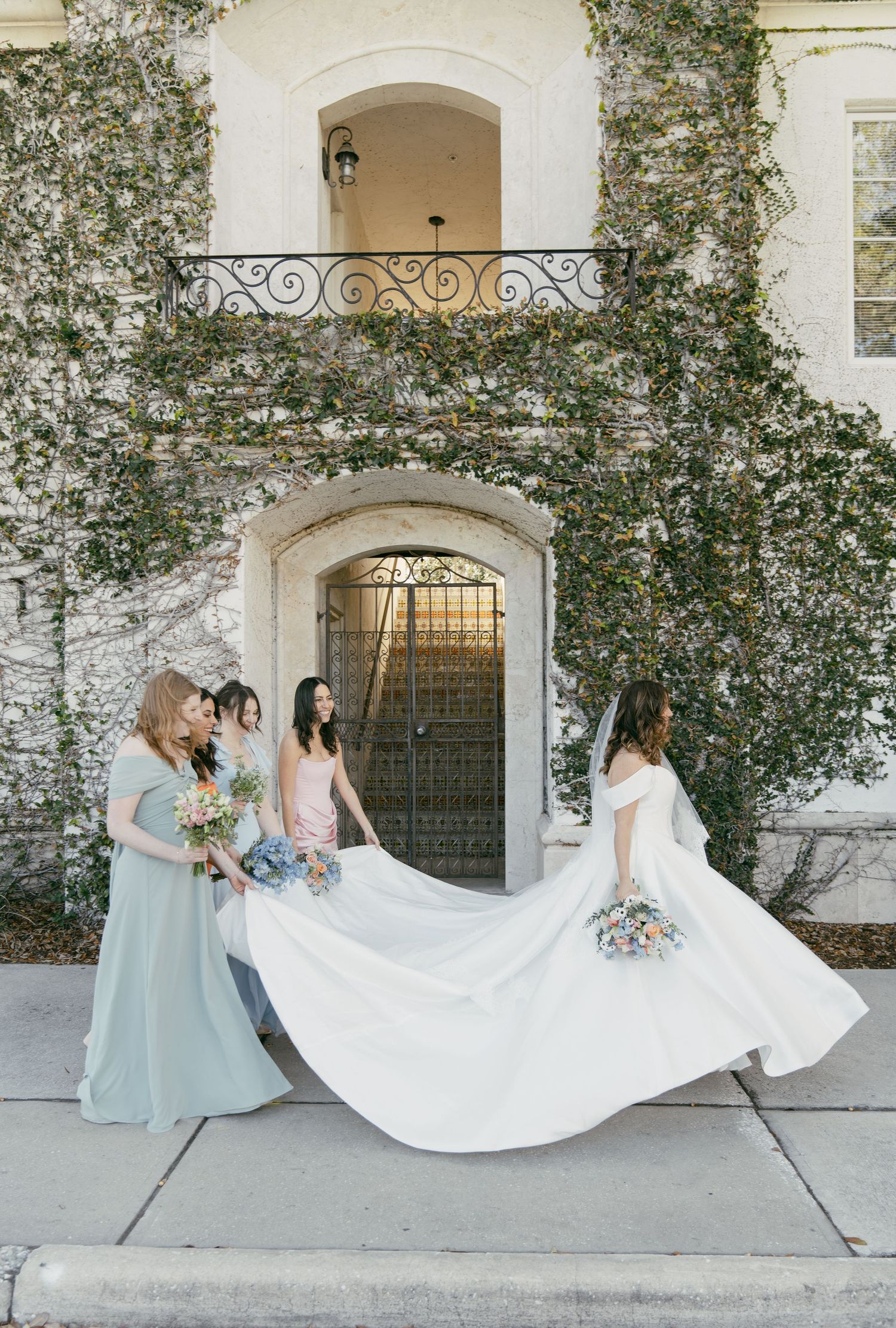 Several bridesmaids in pale blue dresses help adjust the long white train of a wedding gown outside.