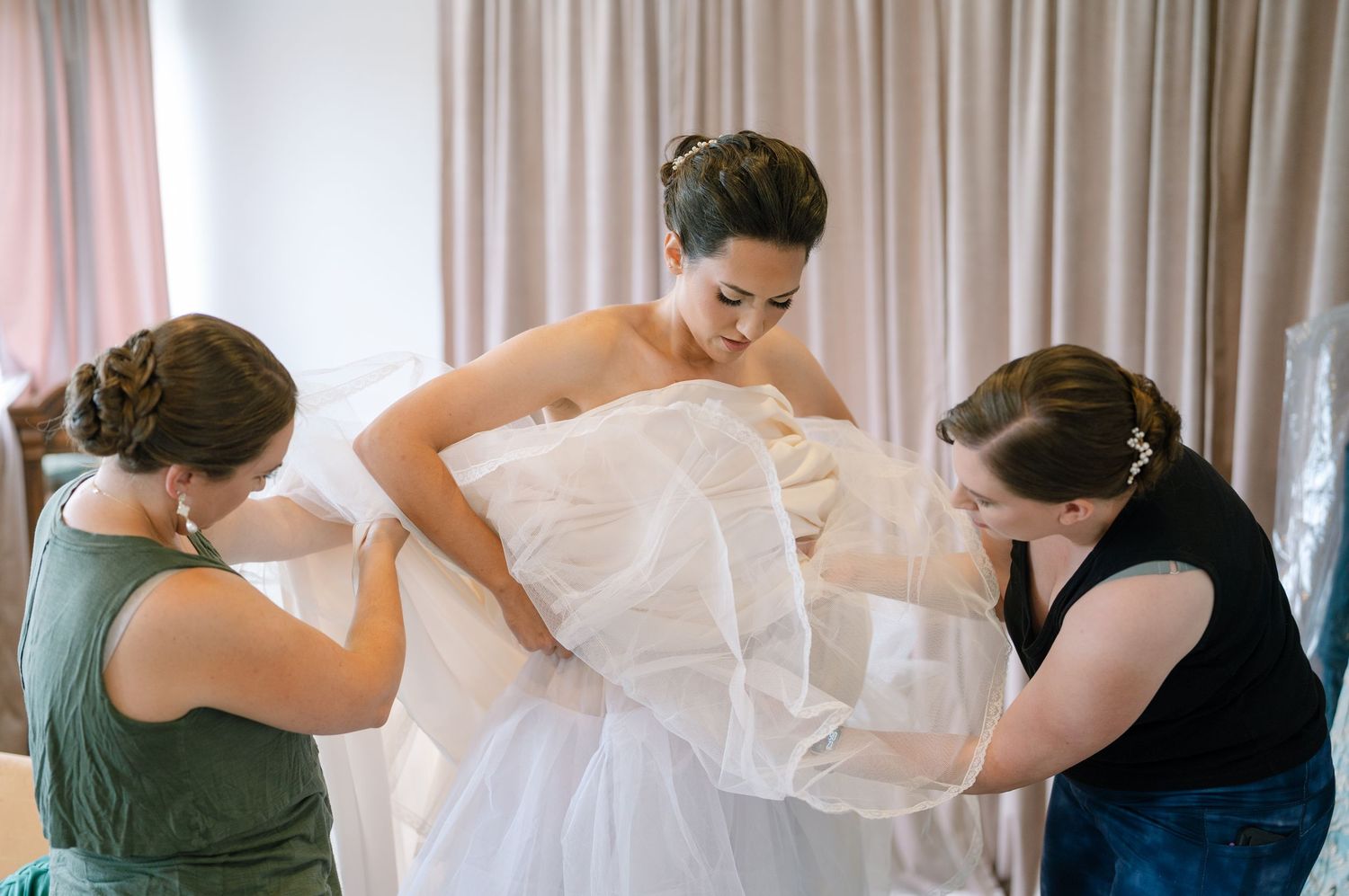 Two assistants help with the final adjustments of a strapless white wedding gown in a bright room.