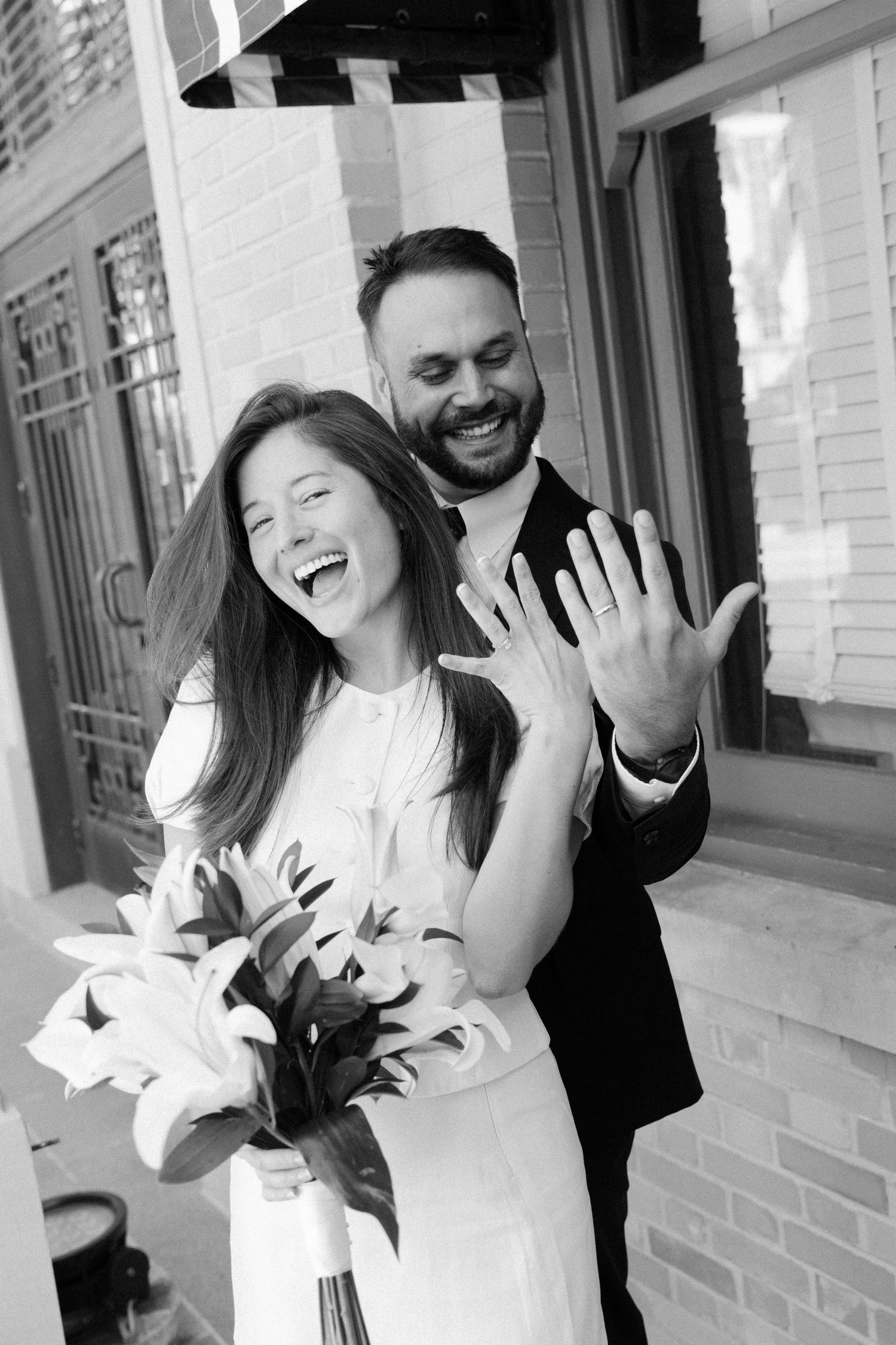 Joyful wedding couple laughing together while holding white lilies in a casual outdoor setting.