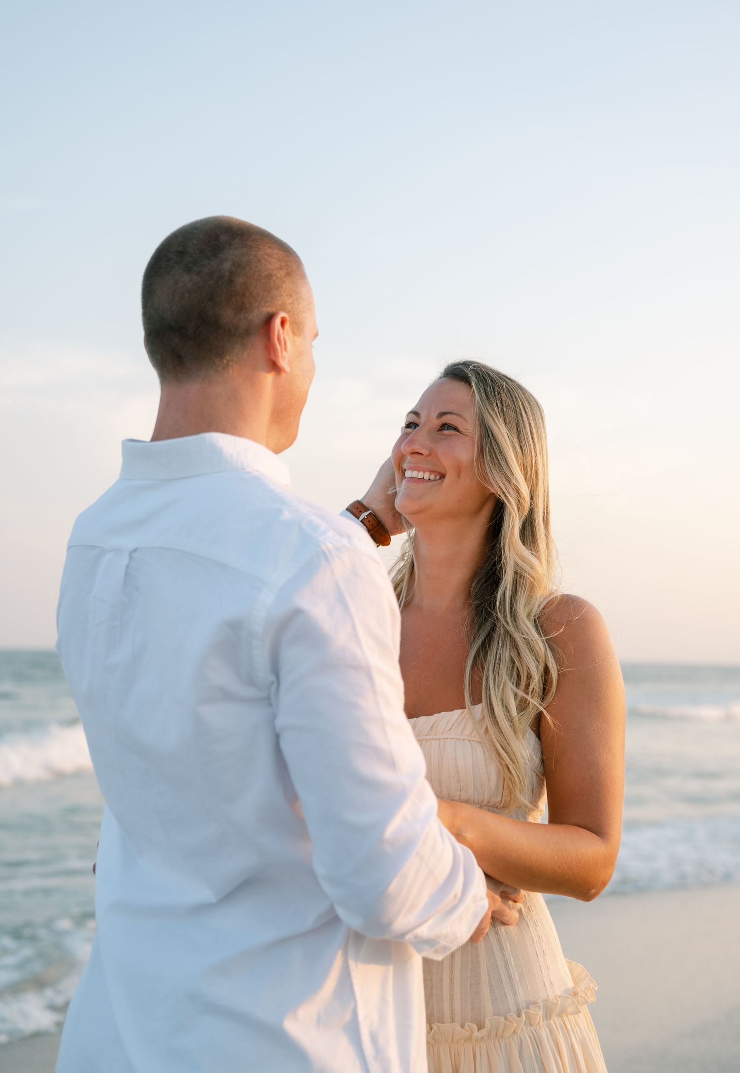 A couple laughs and embraces while standing together on a peaceful beach during golden hour.