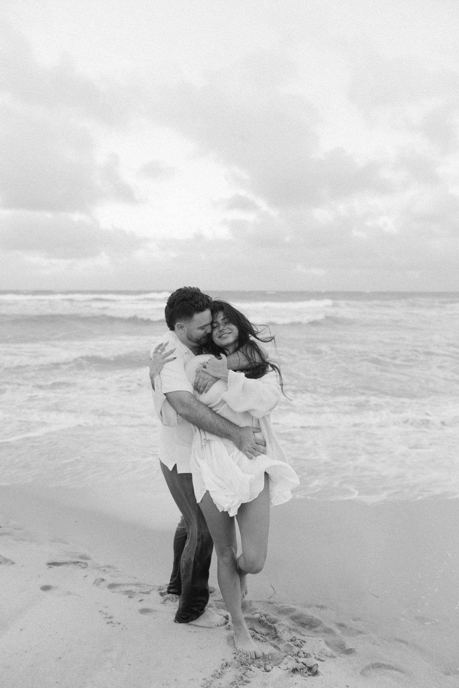A romantic black and white photo series of a couple embracing on a cloudy beach with ocean waves in the background.