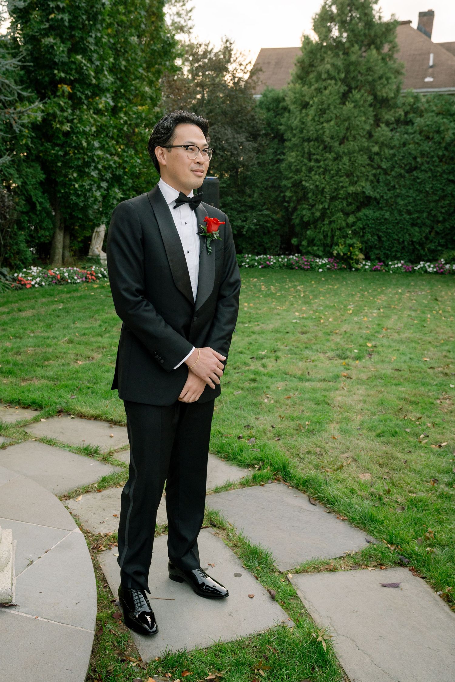 Groom in classic black tuxedo poses on garden pathway during wedding photoshoot.