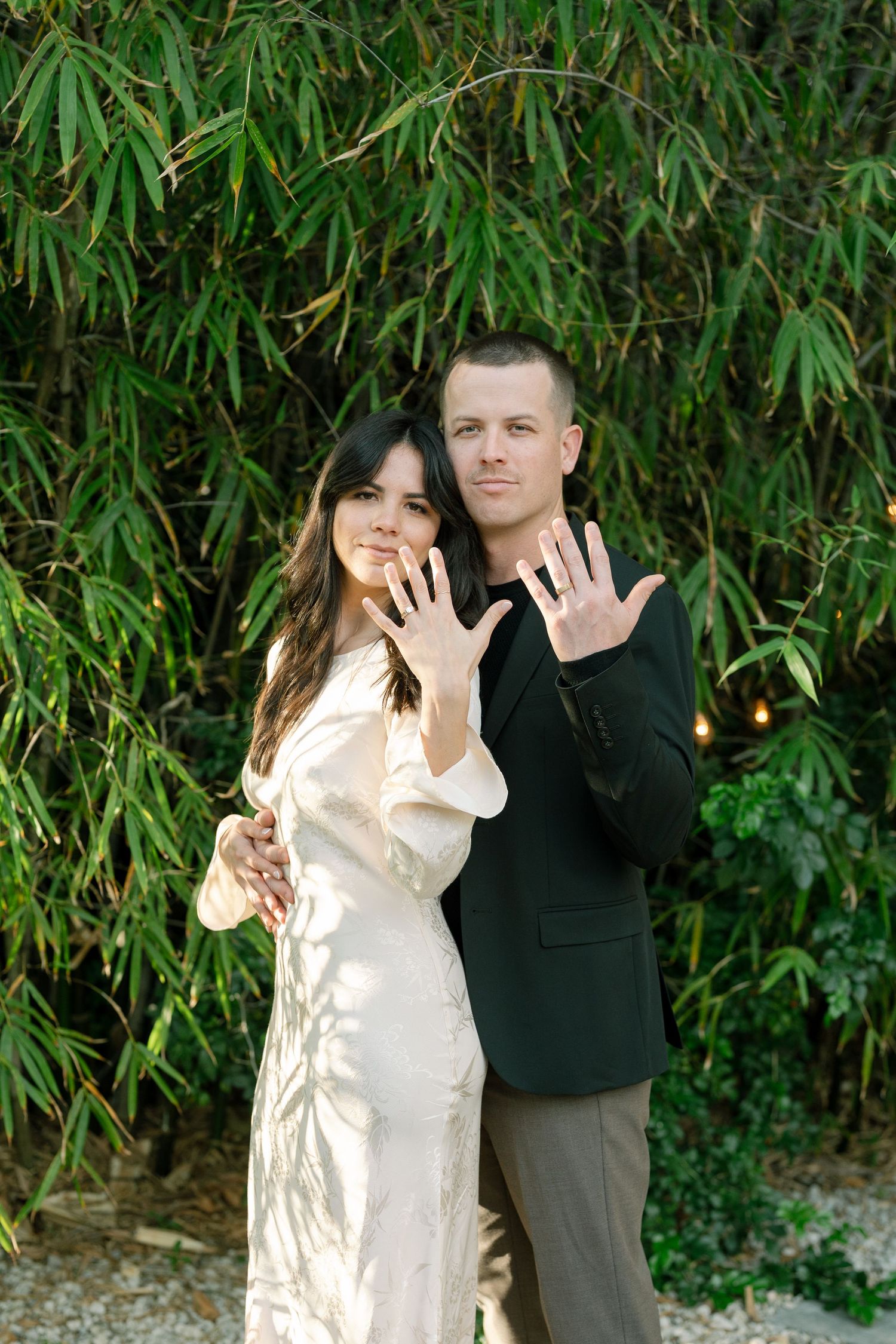 A couple in contrasting outfits showing their hands to the camera against green bamboo foliage.