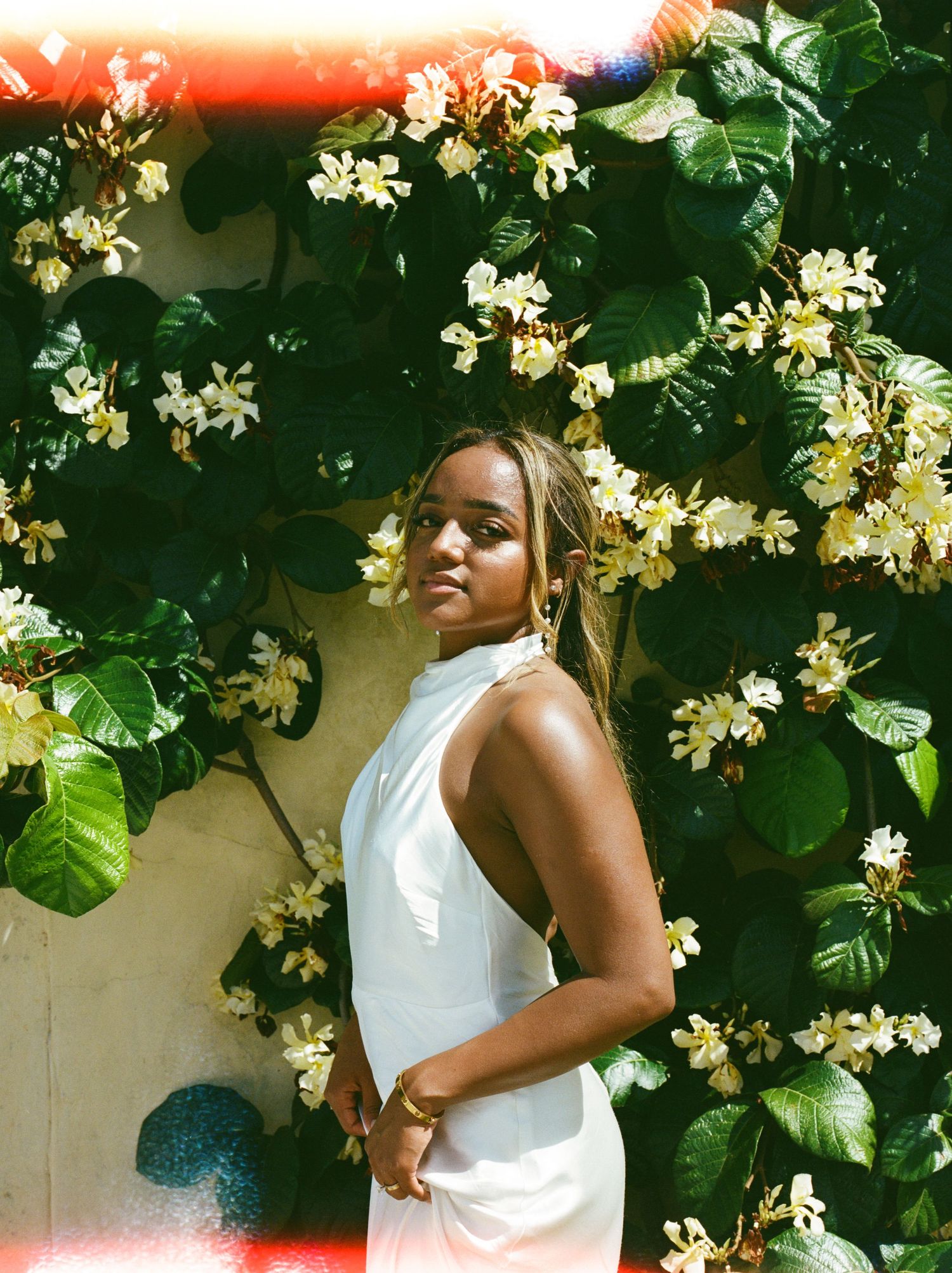 A person in a white dress poses gracefully among blooming white flowers and lush green foliage in a garden setting.