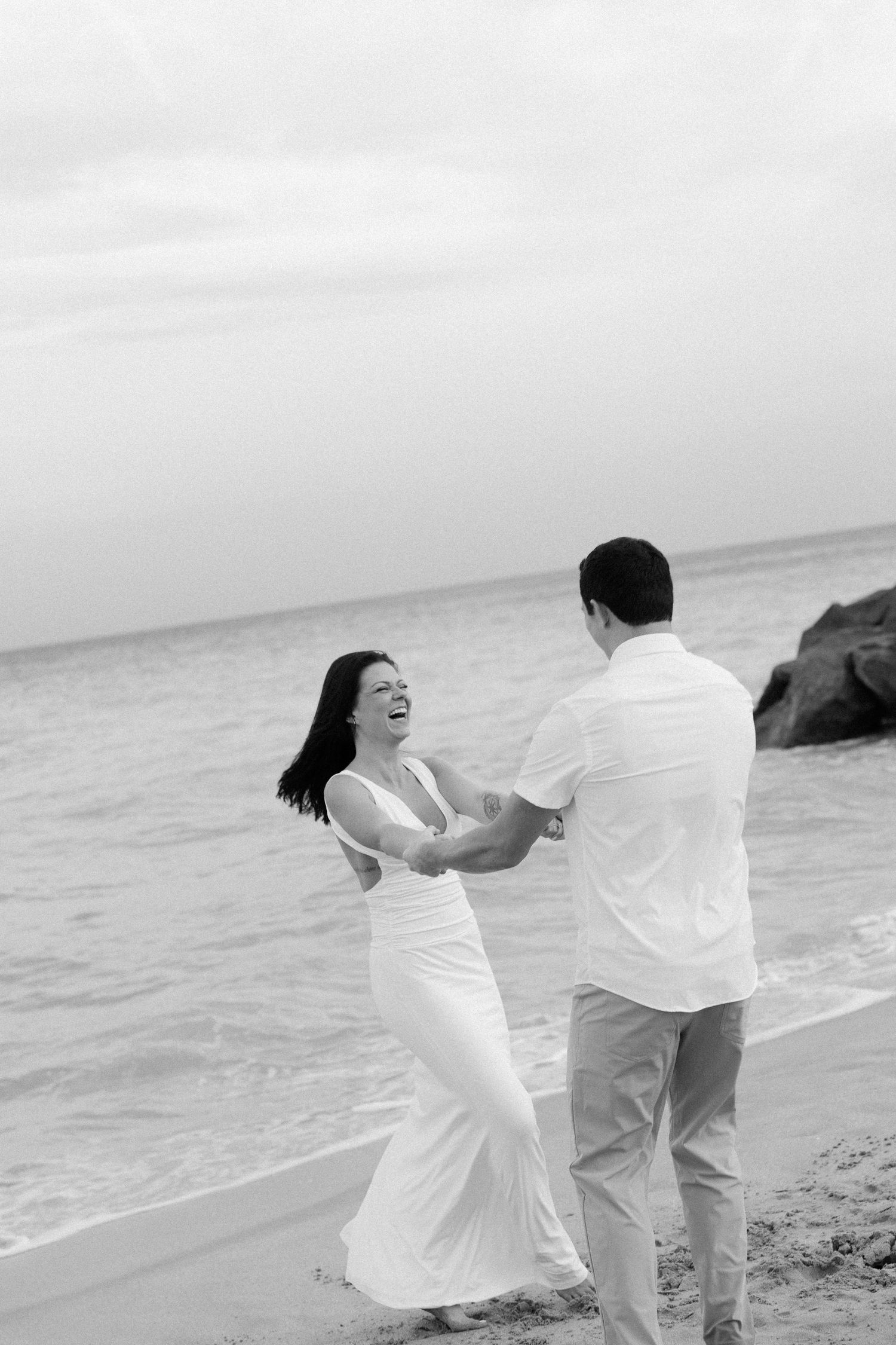 Dancing couple in white clothing twirl playfully on beach shore during overcast day.