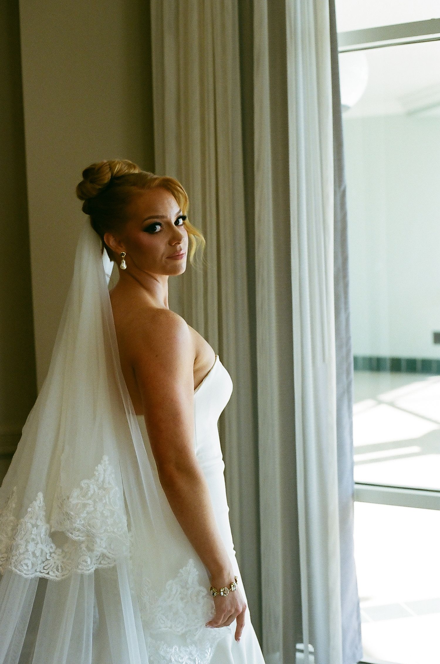A bride in a white strapless wedding gown poses elegantly near a window, showing the dress's beautiful silhouette.