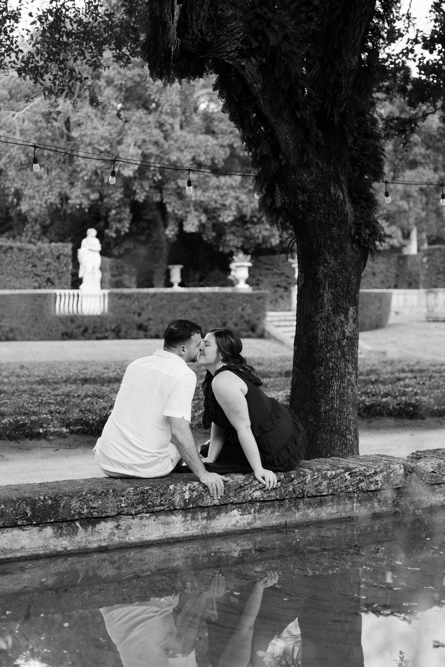 A couple sits together by a reflective pond in a peaceful garden setting, creating a romantic black and white scene.