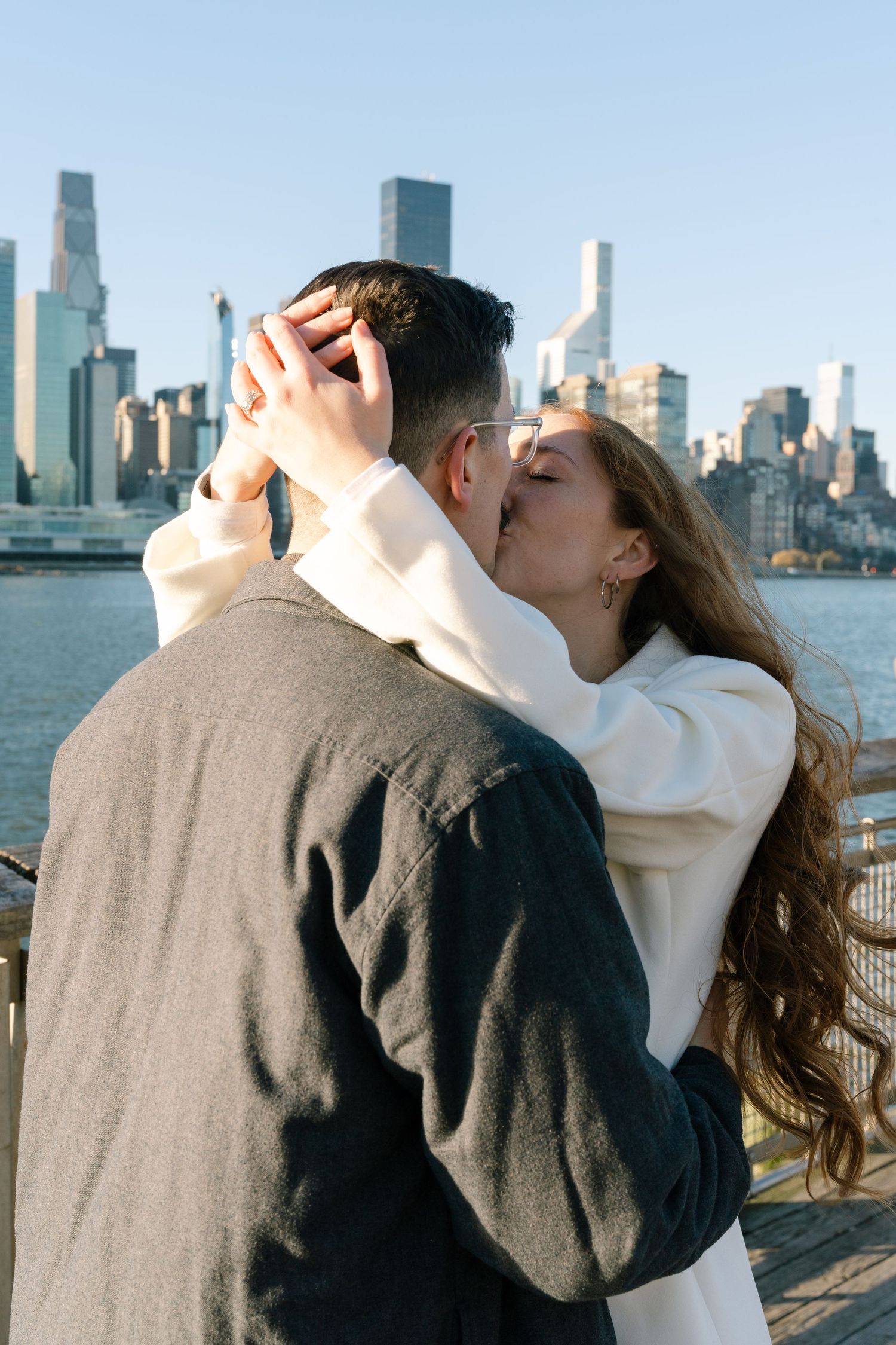 A romantic couple sharing a kiss against the New York City skyline backdrop on a sunny day.