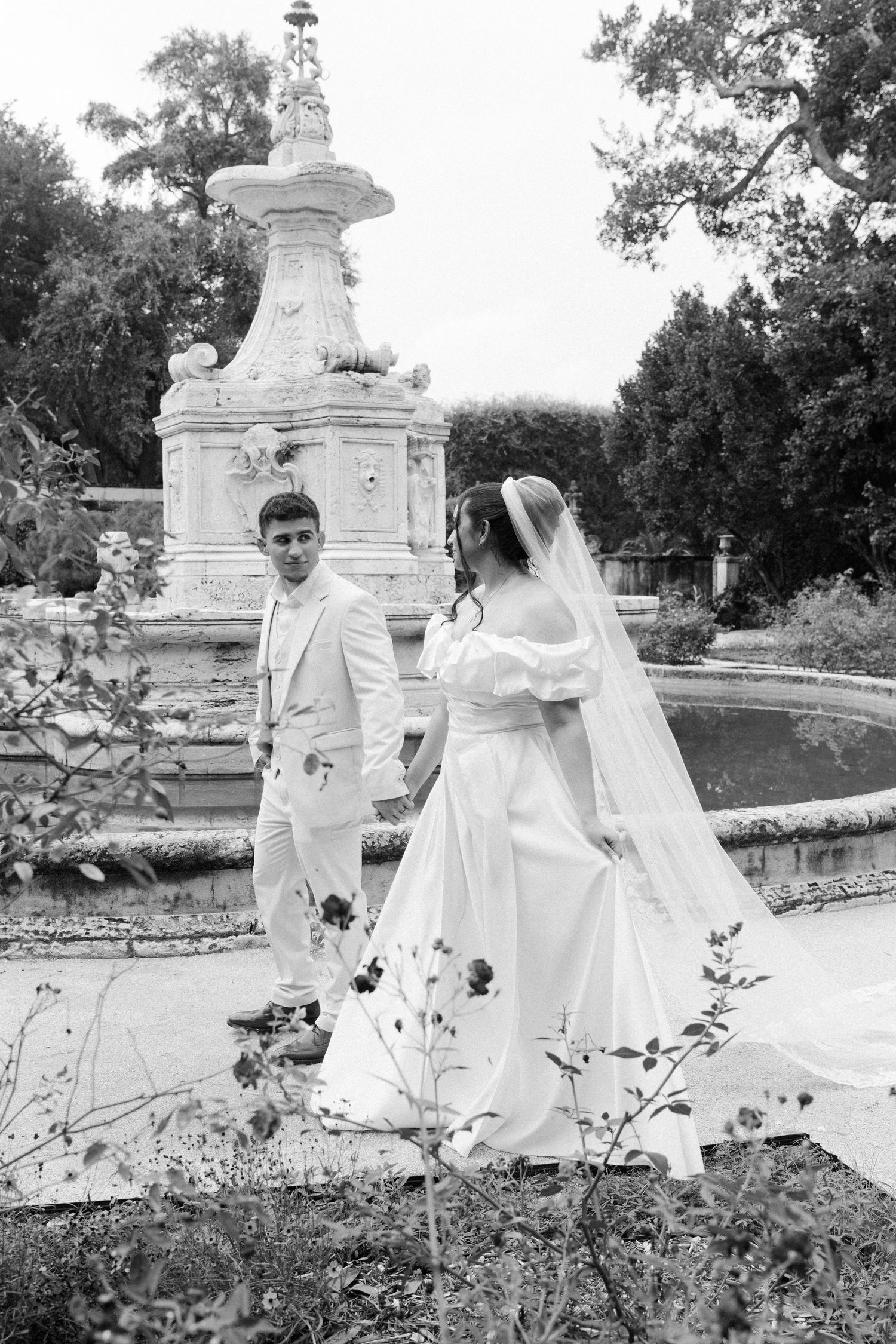 A couple in white wedding attire stands beside an ornate stone fountain in a garden setting, black and white photograph.