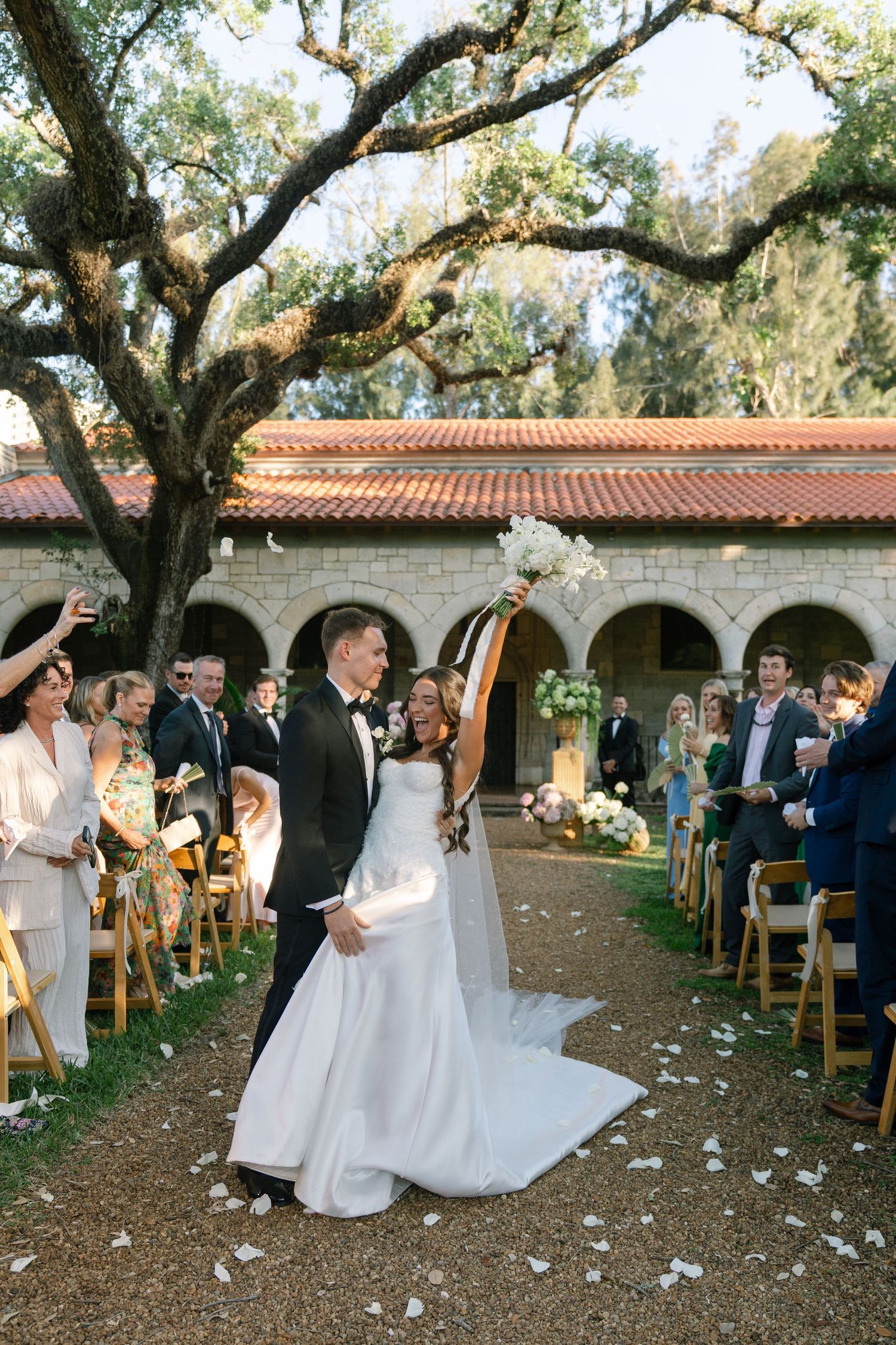Bride and groom walk down outdoor aisle after wedding ceremony under oak tree at elegant venue.