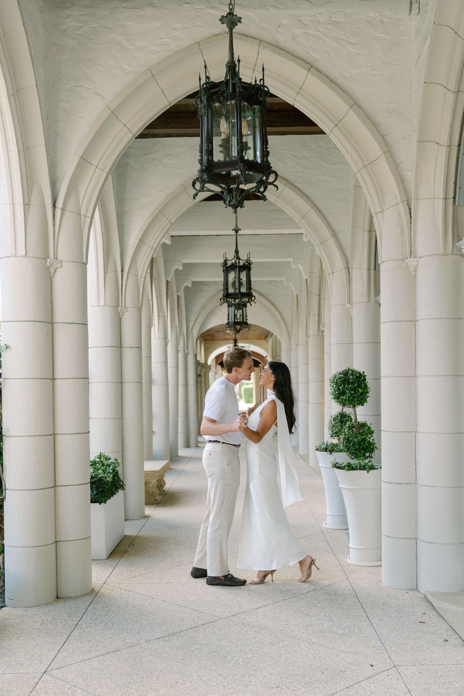 Couple sharing a romantic moment in an elegant white arched colonnade with hanging lanterns and topiary plants.