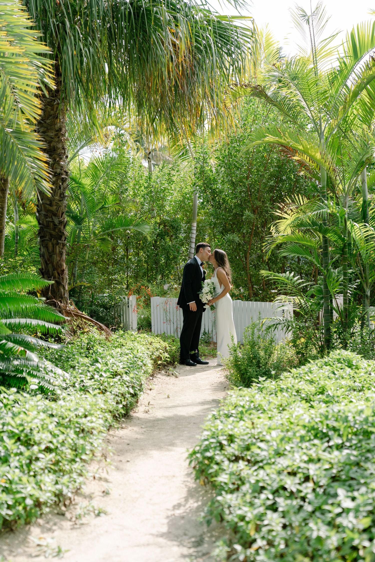 Bride and groom share a romantic moment on a lush tropical garden path lined with greenery and palm trees.