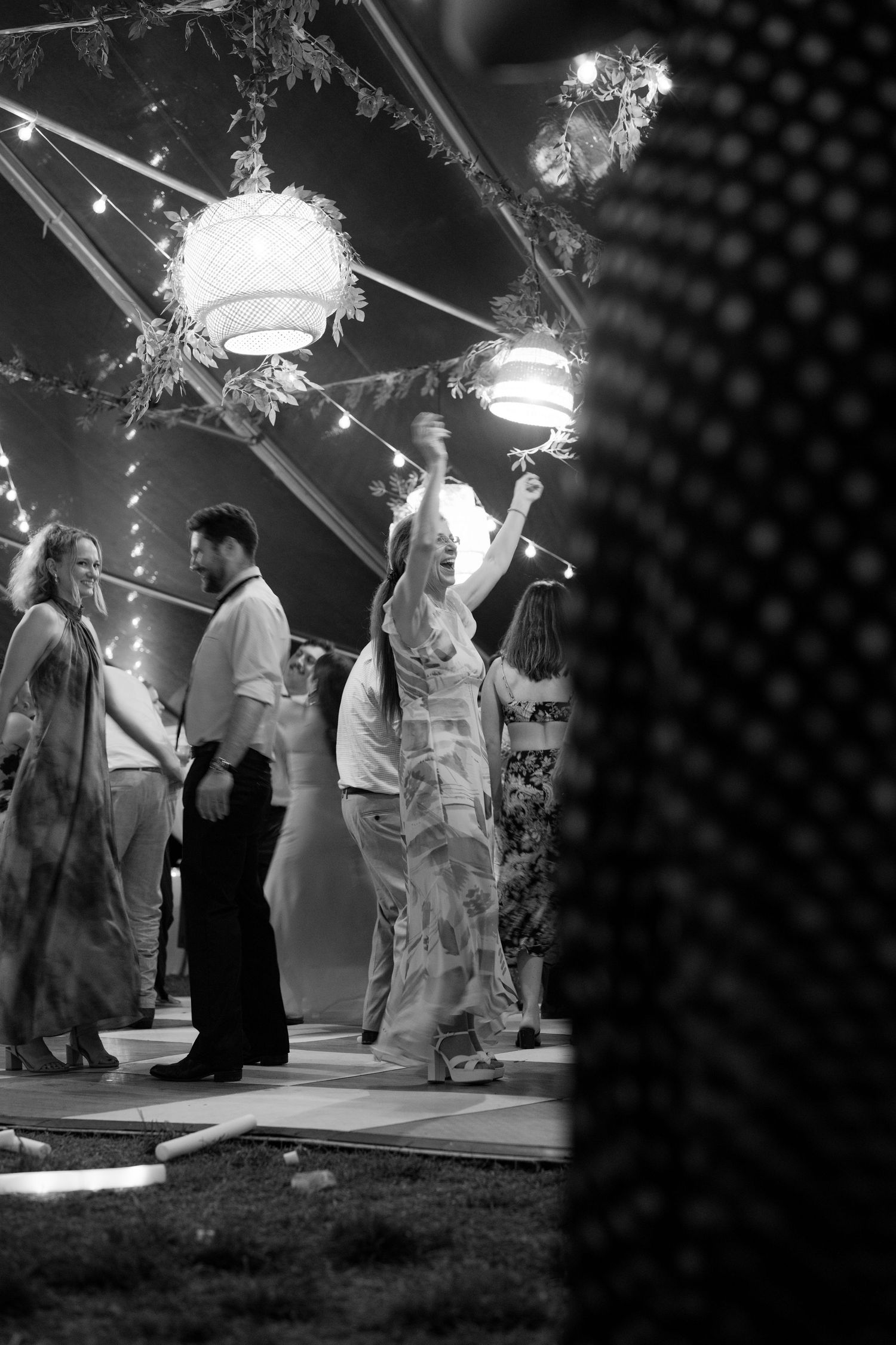 Black and white photo of wedding guests dancing under string lights and lanterns in outdoor tent at night.