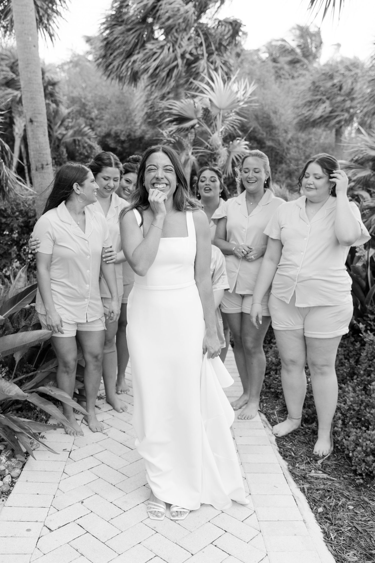 Black and white photo of a bride in a sleek white gown smiling with bridesmaids in casual pajamas outdoors.