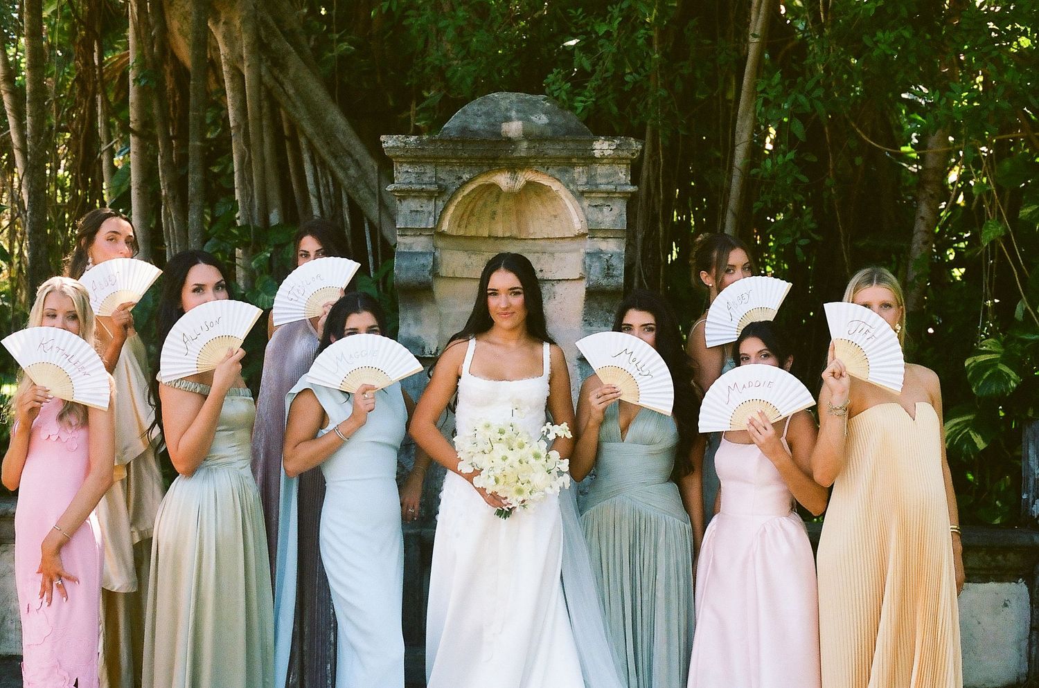 Bride in white gown holds bouquet surrounded by bridesmaids in pastel dresses holding white fans outdoors.