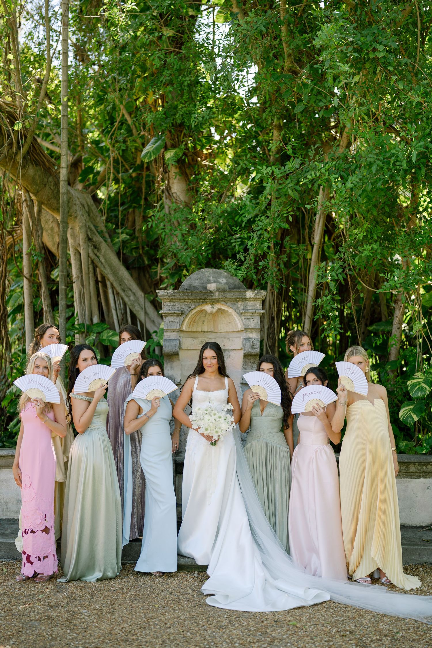 Bride in white gown poses with bridesmaids in mismatched pastel dresses holding white fans in lush garden.
