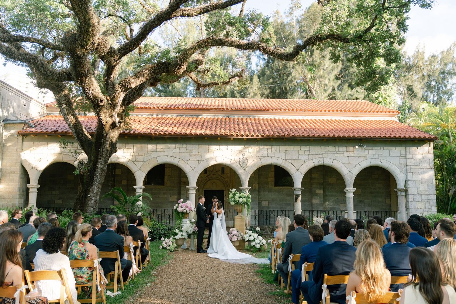 Outdoor wedding ceremony under oak tree at stone arched chapel with seated guests.