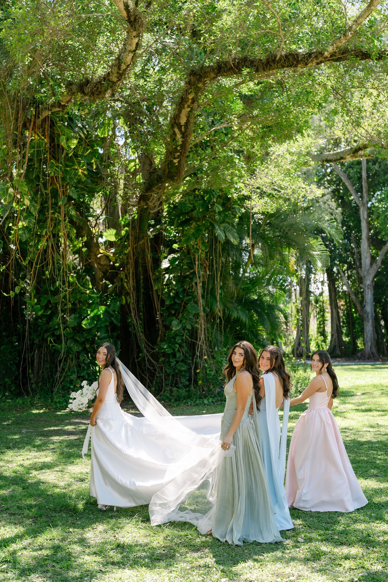 A bride in a white gown and bridesmaids in sage and blush dresses pose under a large banyan tree.