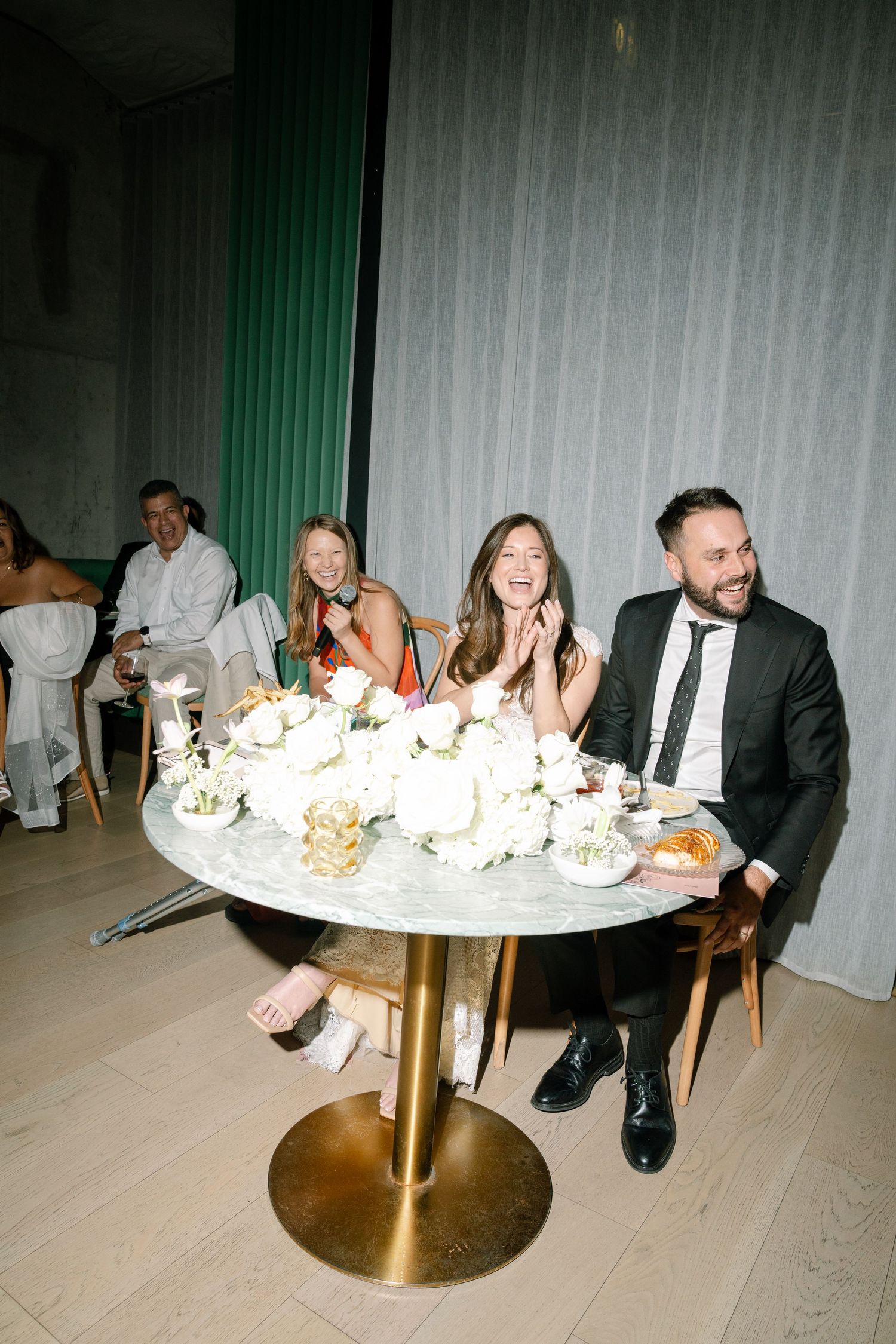 Wedding guests seated at an elegant round table with white floral centerpieces and gold accents.