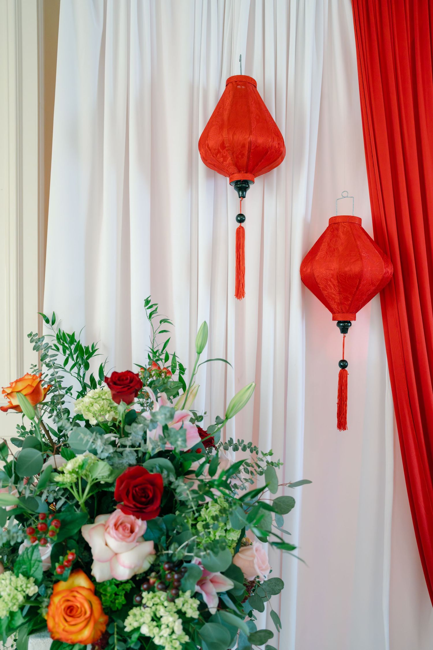 Wedding decor featuring red lanterns and floral arrangements with roses against white and red drapery.