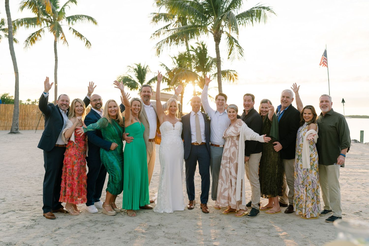 Wedding guests gathered on a beach with palm trees at sunset.