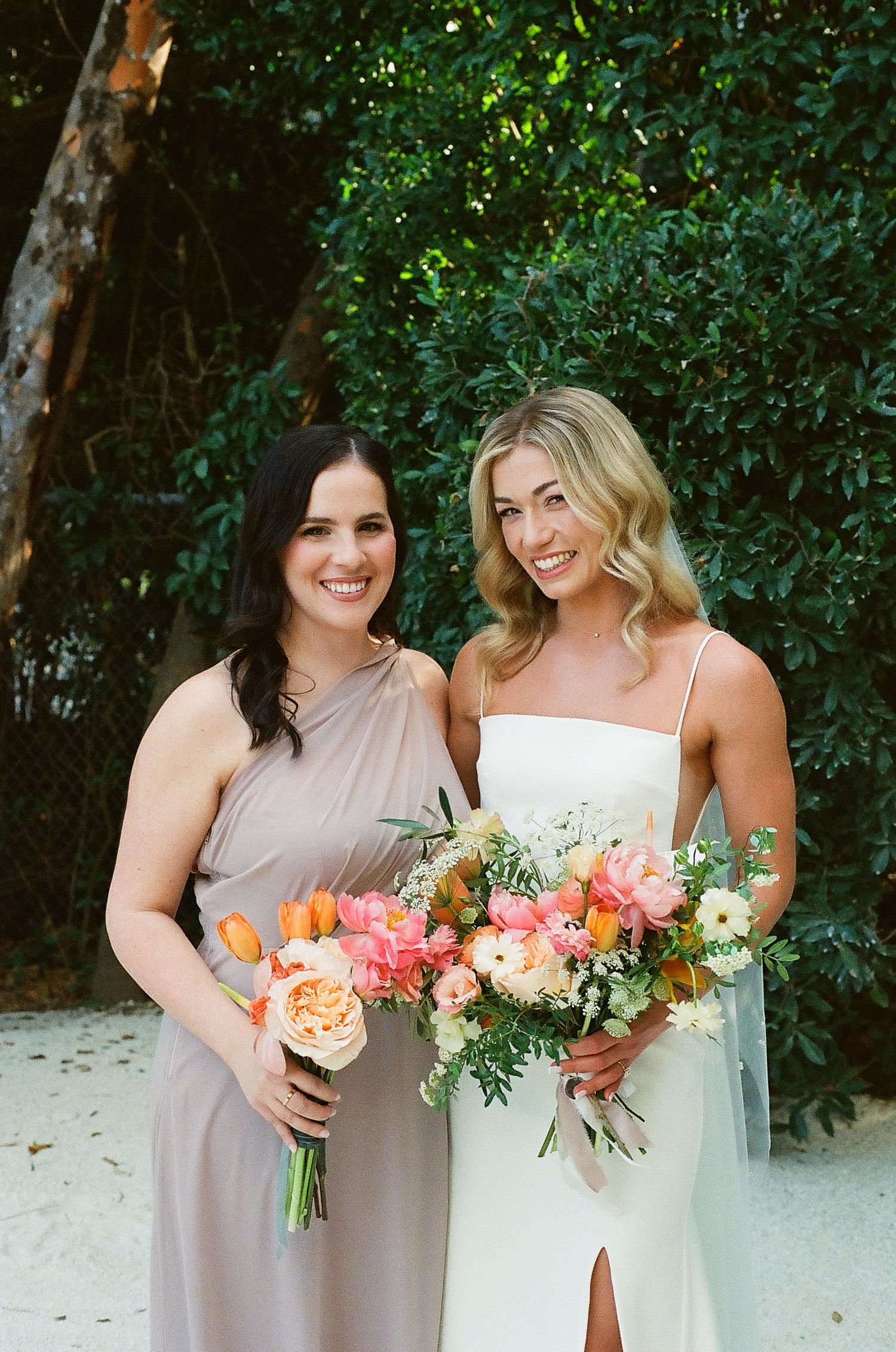 Wedding party members pose with coral and peach bouquets against green foliage.