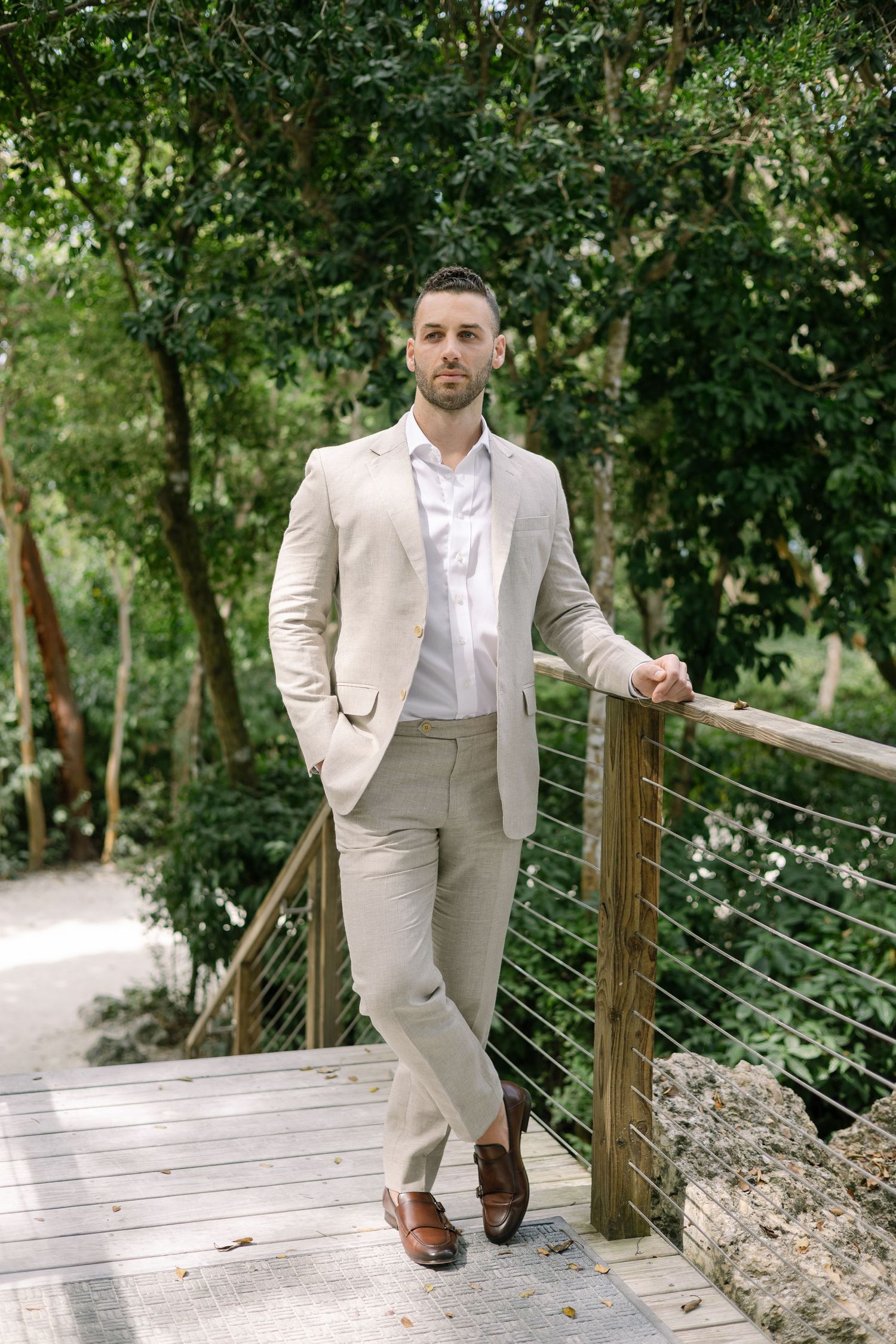 Man in light beige suit walks along wooden deck pathway surrounded by lush greenery.