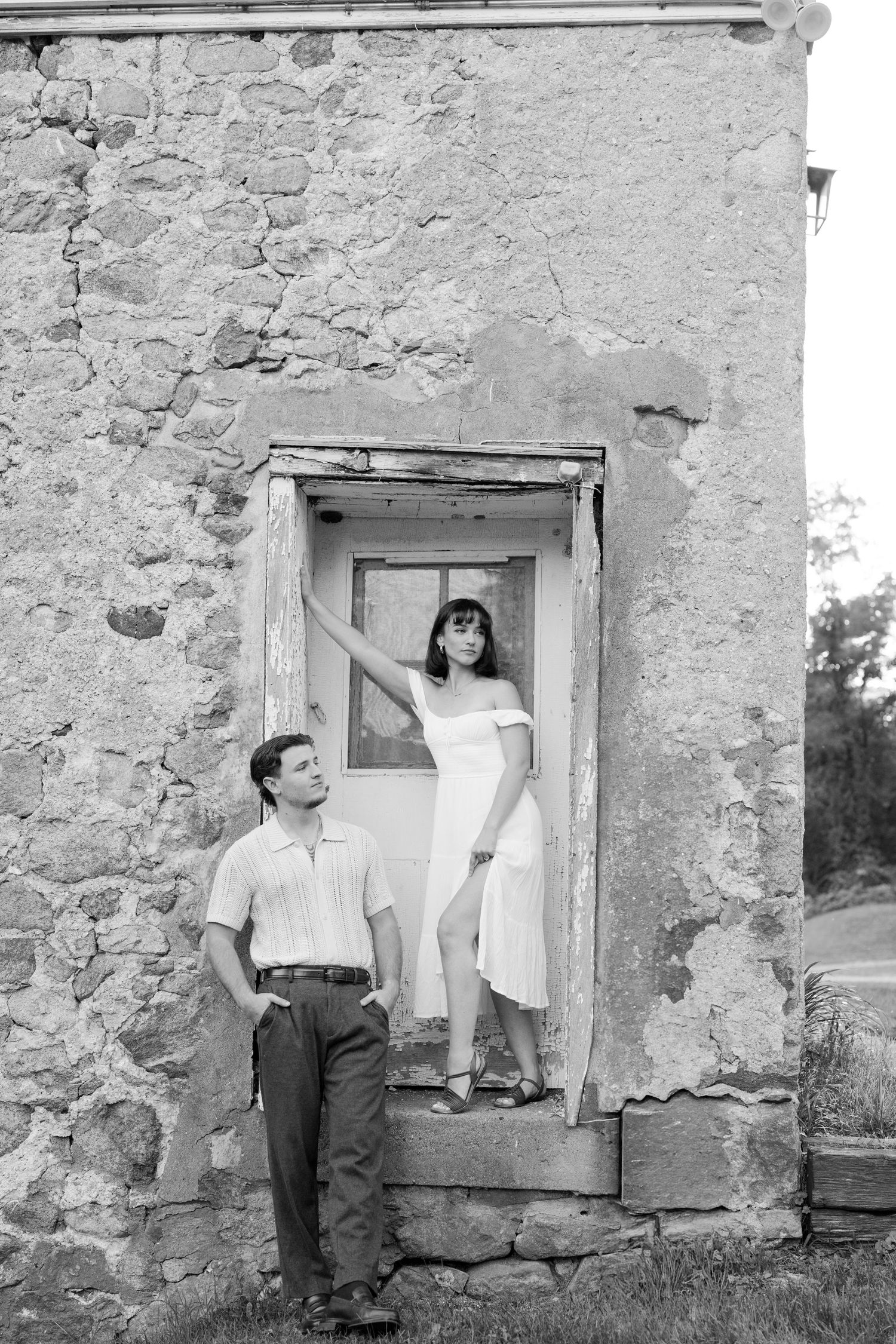 Black and white photo of a couple posing by a rustic stone building, woman in white dress standing in doorway.