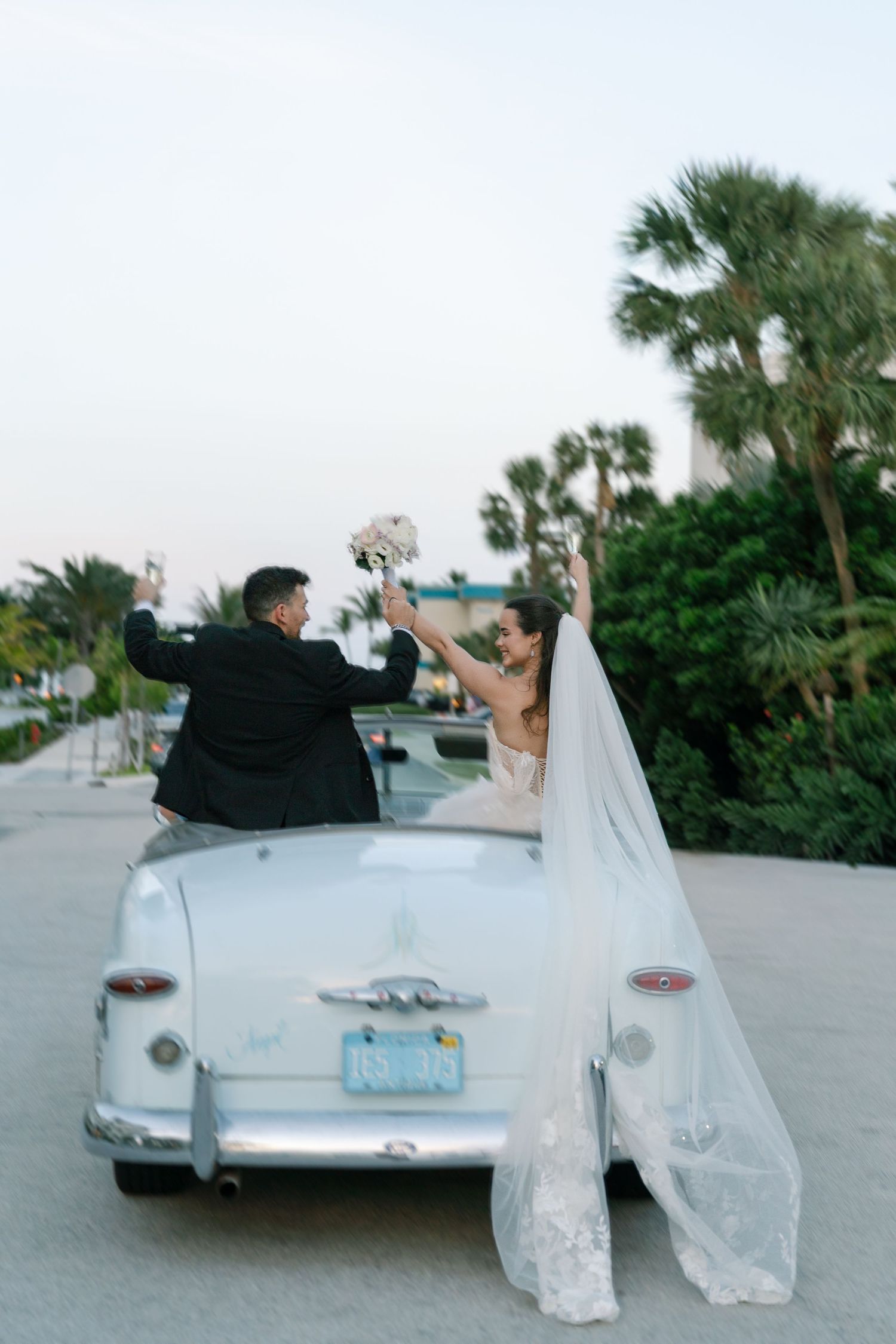 Bride and groom celebrating beside a vintage white convertible car, with palm trees in the background.