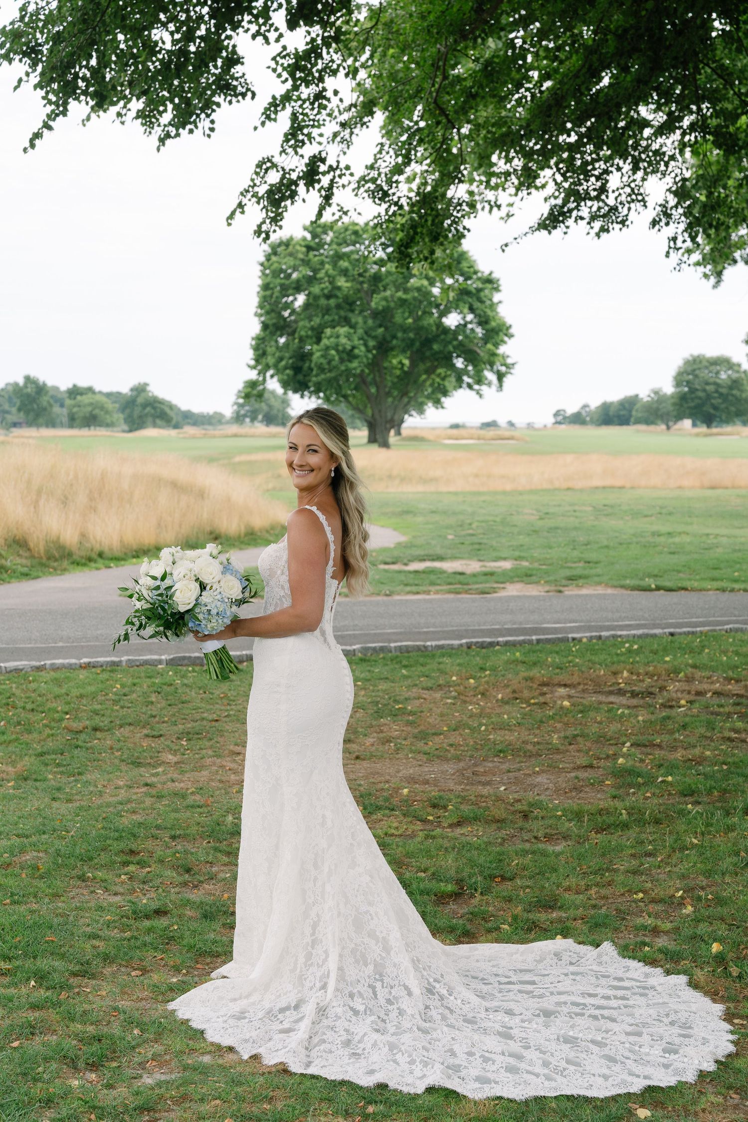 Bride in elegant lace mermaid wedding gown with train holds white bouquet on green lawn under large tree.