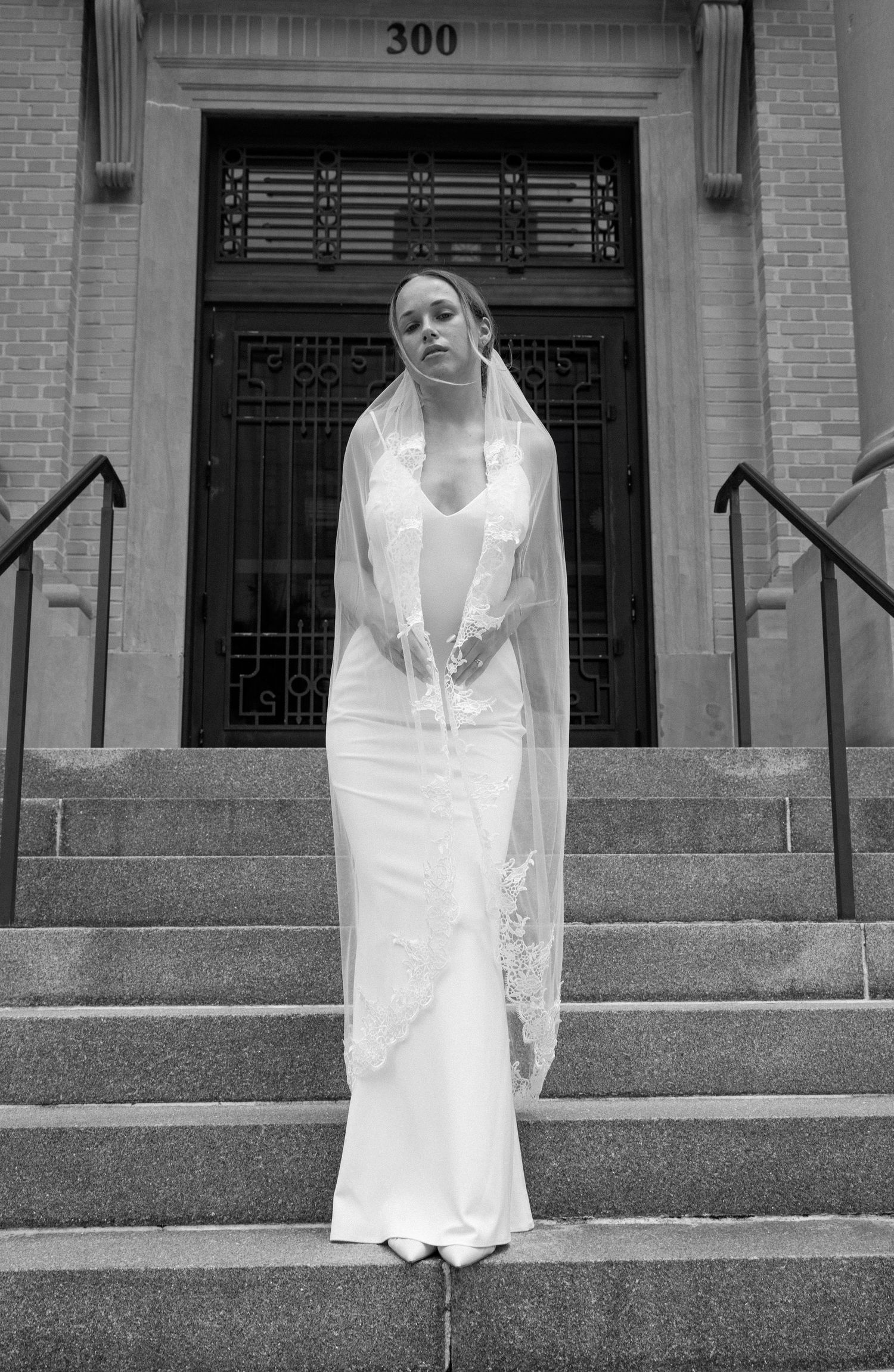 A person in a flowing white dress sits on outdoor stone steps in front of a brownstone building entrance.