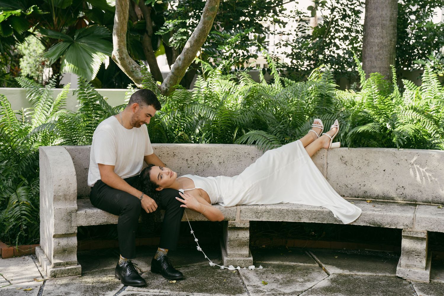 A couple relaxes together on a stone bench in a lush garden setting on a sunny day.