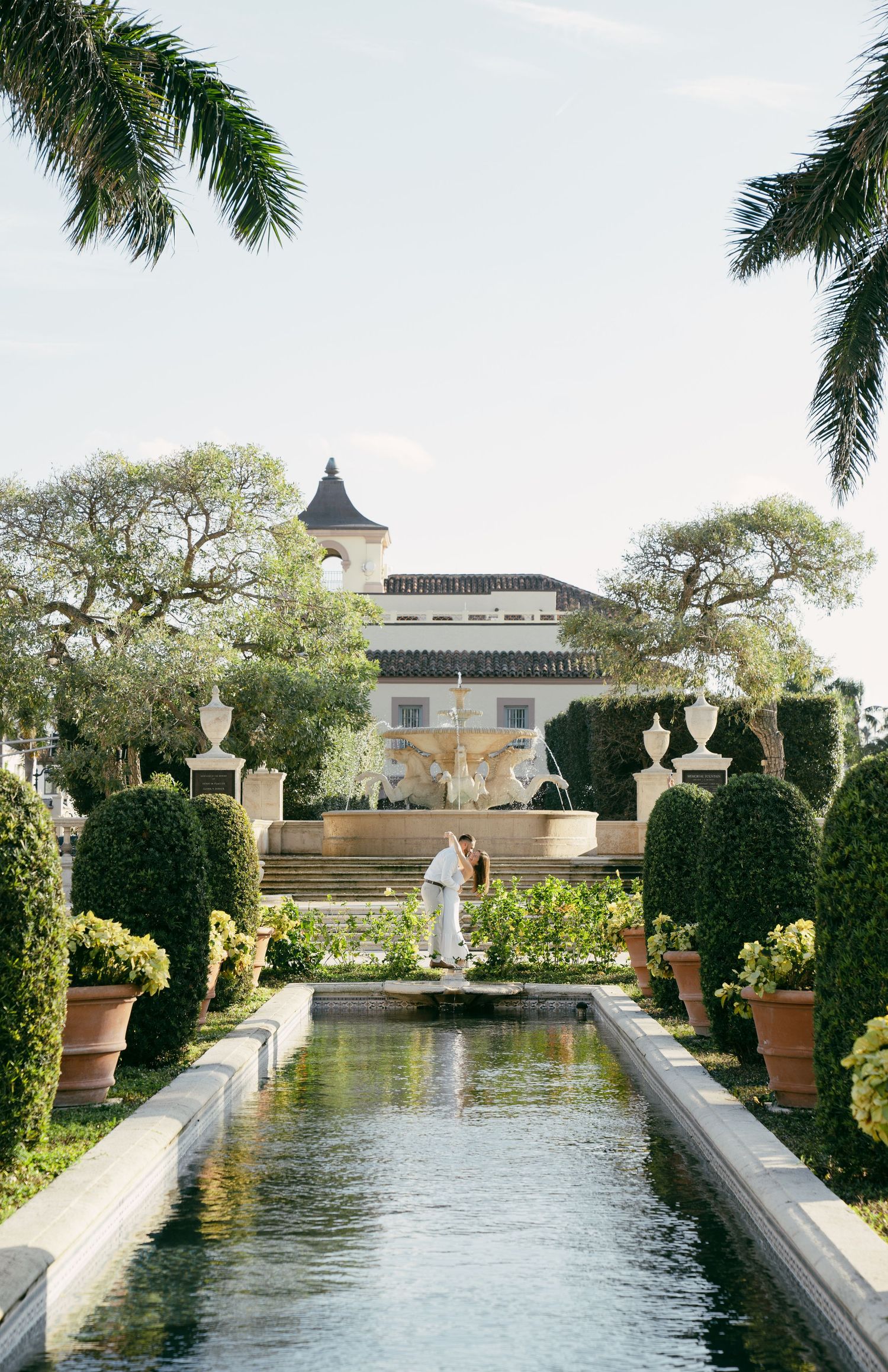 Elegant garden courtyard features a long reflecting pool flanked by manicured topiaries and palm trees leading to a white mansion.