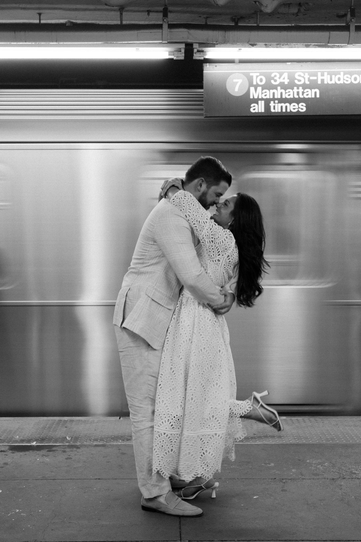 Black and white photo of a couple embracing on a NYC subway platform as a train blurs behind them.