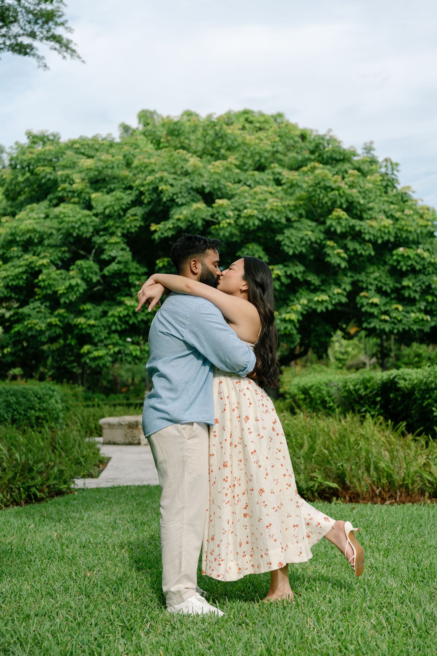 Couple embracing and kissing in a lush green garden, woman in floral skirt, man in blue shirt.