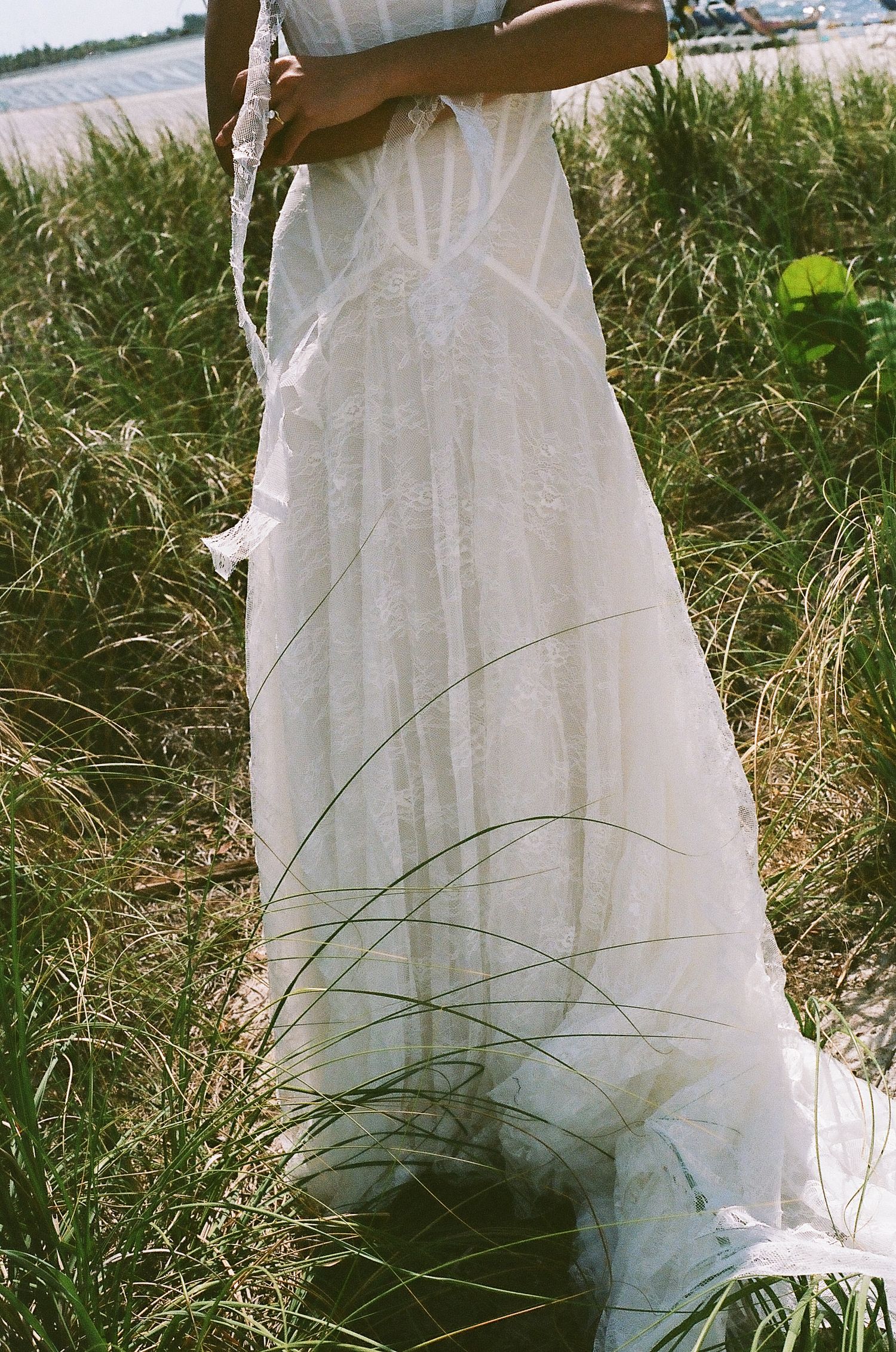 Person in white lace wedding gown standing in tall grass near a beach, holding a floral accessory.