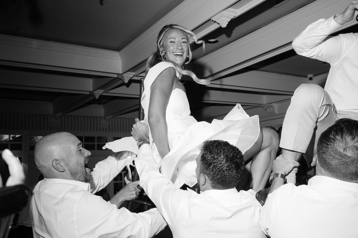 Bride being lifted by groomsmen during joyful wedding reception celebration in black and white photo.
