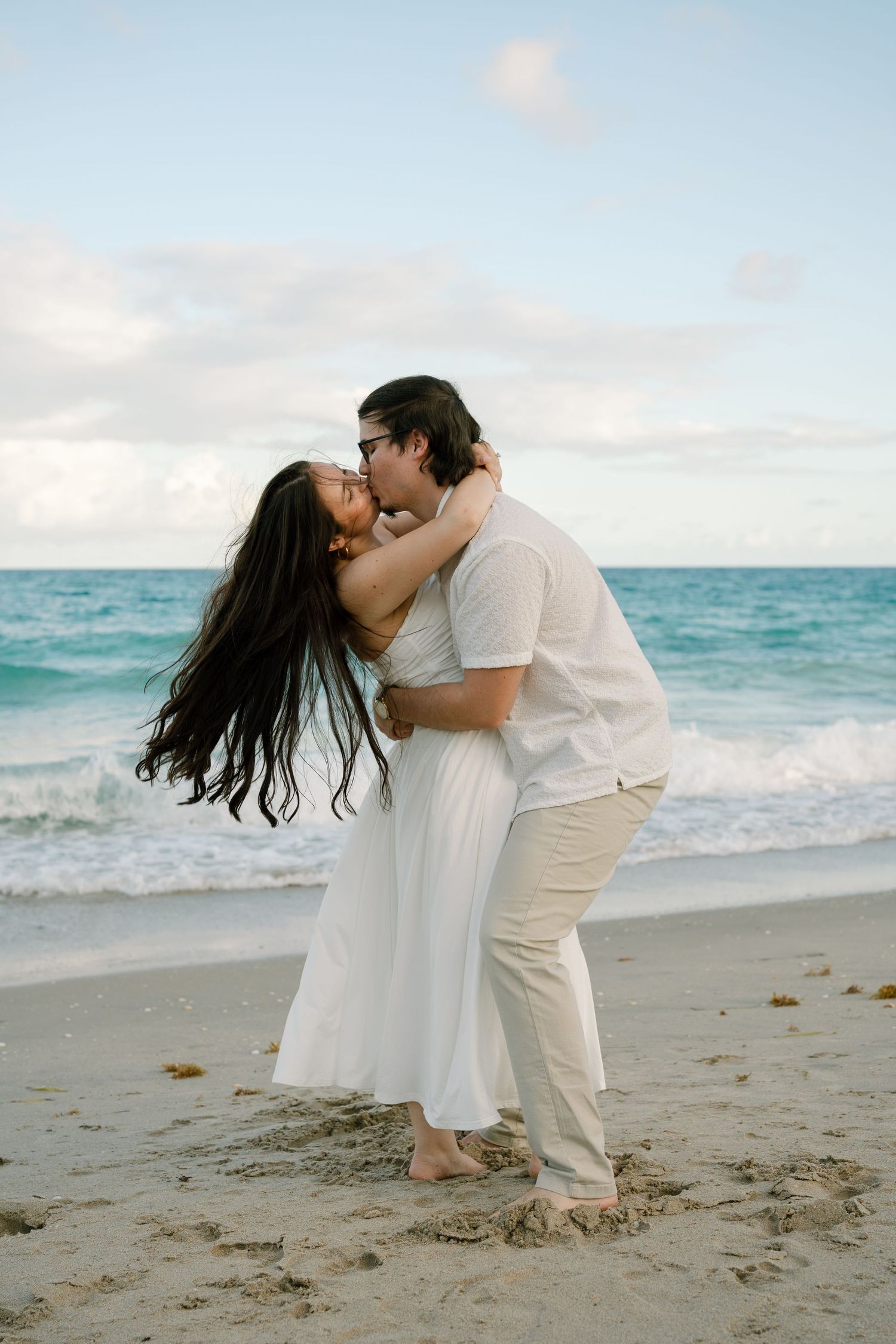 A couple in white clothing shares a romantic embrace on a sandy beach with turquoise ocean waves.