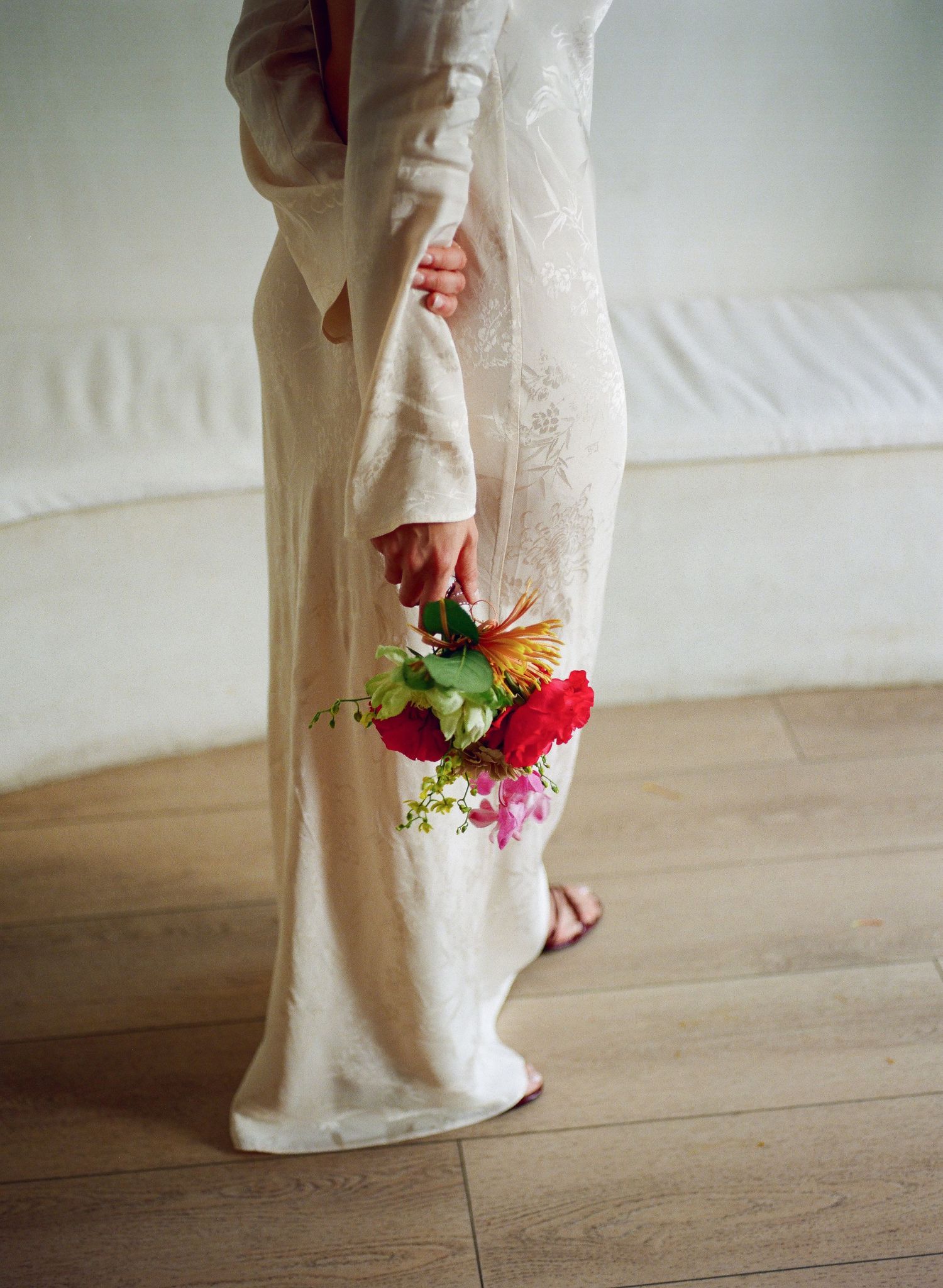 Person in white clothing holding a vibrant bouquet of red and pink flowers against wooden flooring.