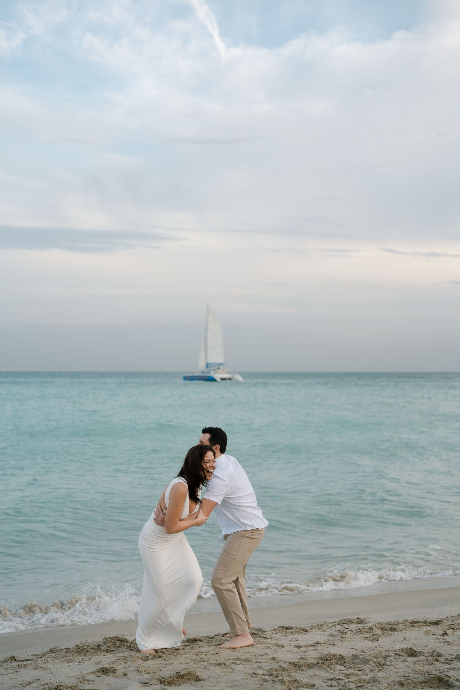 A romantic couple shares a tender moment on a beach with turquoise waters and a sailboat in the distance.
