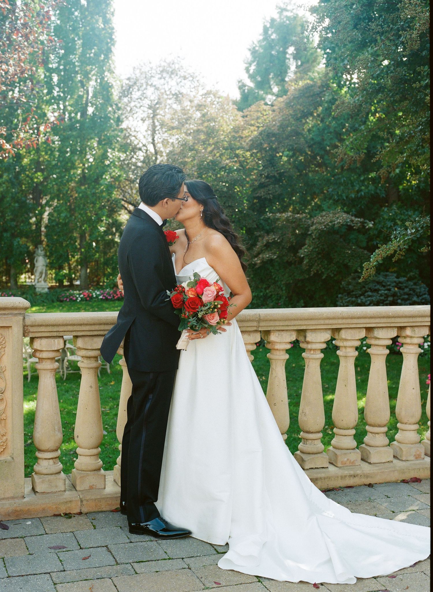 A romantic wedding couple shares an intimate moment on a stone balcony surrounded by lush greenery in a garden setting.
