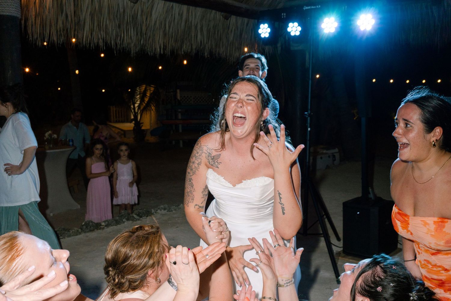 Bride laughing joyfully while being lifted by guests during lively outdoor wedding reception dance party at night.