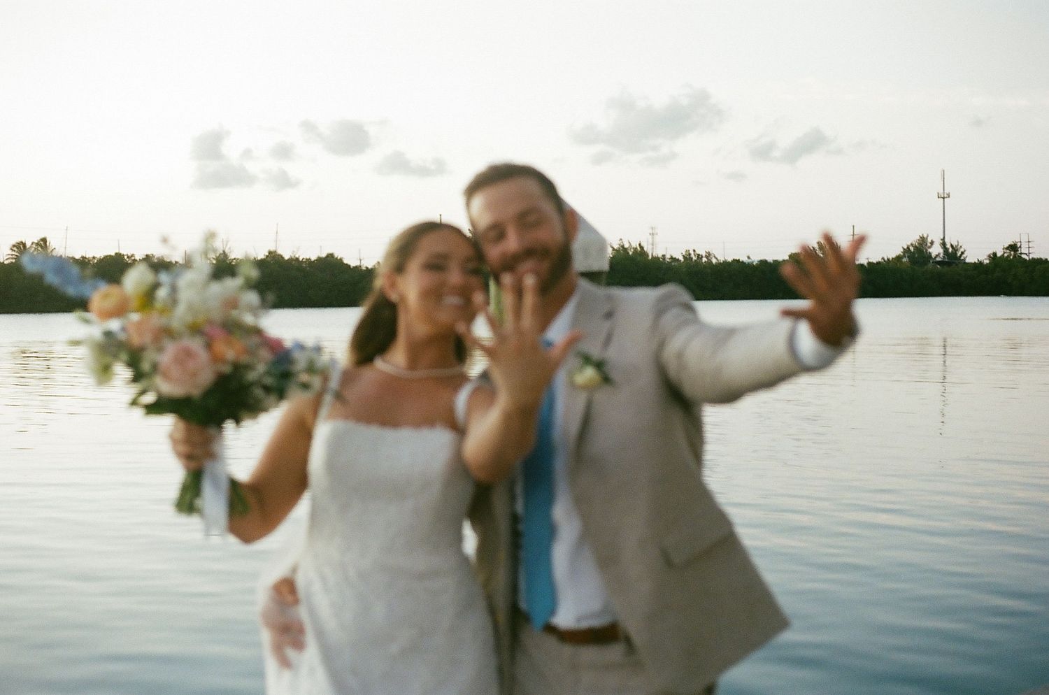 Newlyweds posing joyfully by the water at sunset with bouquet in hand.
