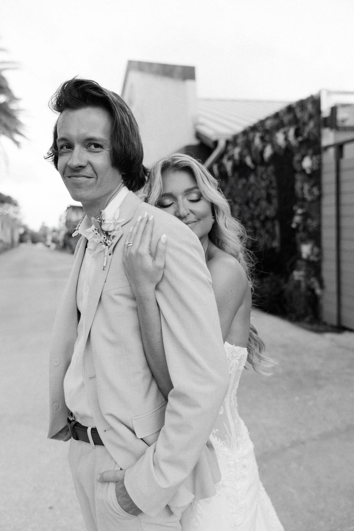 A vintage black and white wedding photograph shows a joyful couple in white attire embracing outdoors.