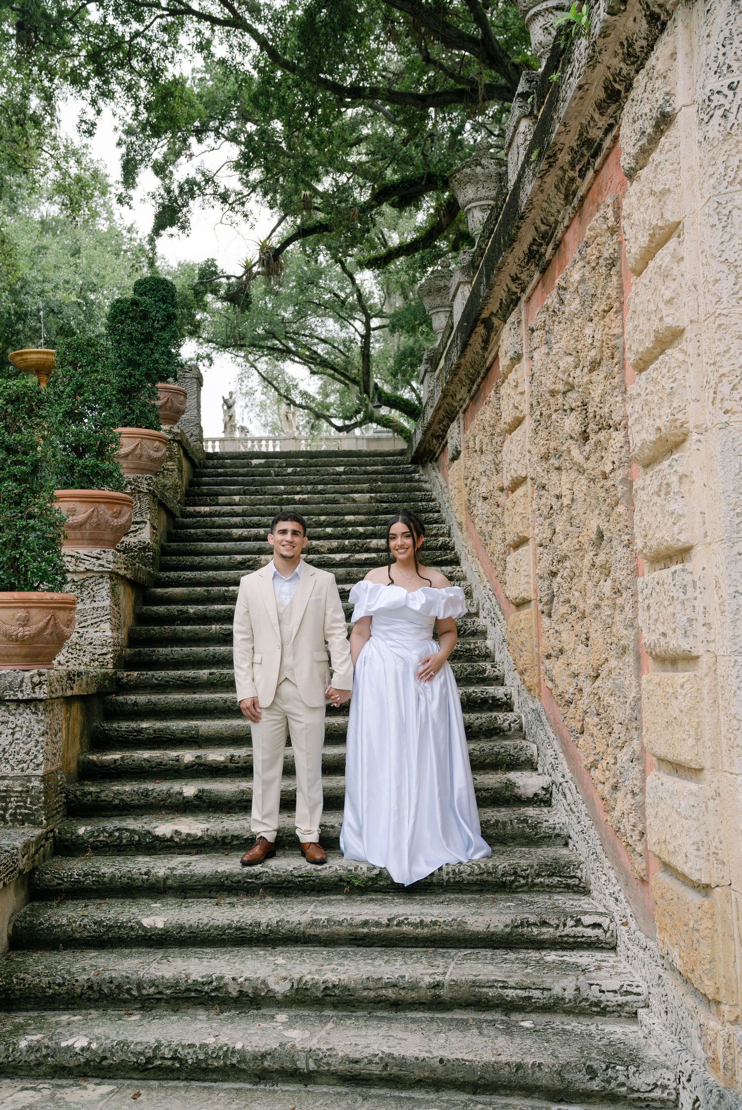 A couple sits on limestone steps surrounded by lush greenery in a romantic garden setting at a historic estate.