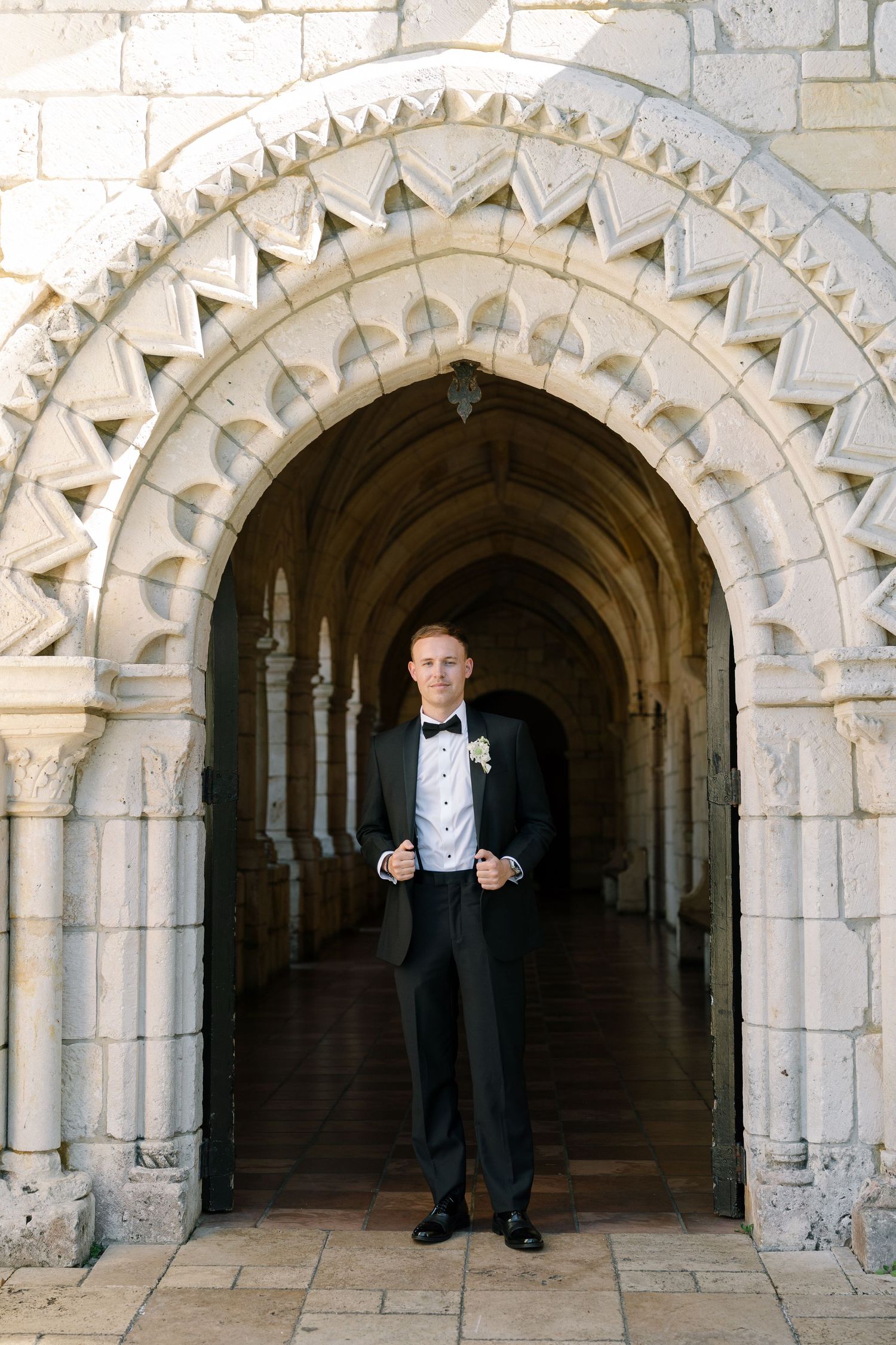 Elegant groom in black tuxedo stands beneath ornate stone Gothic arch at historic wedding venue.