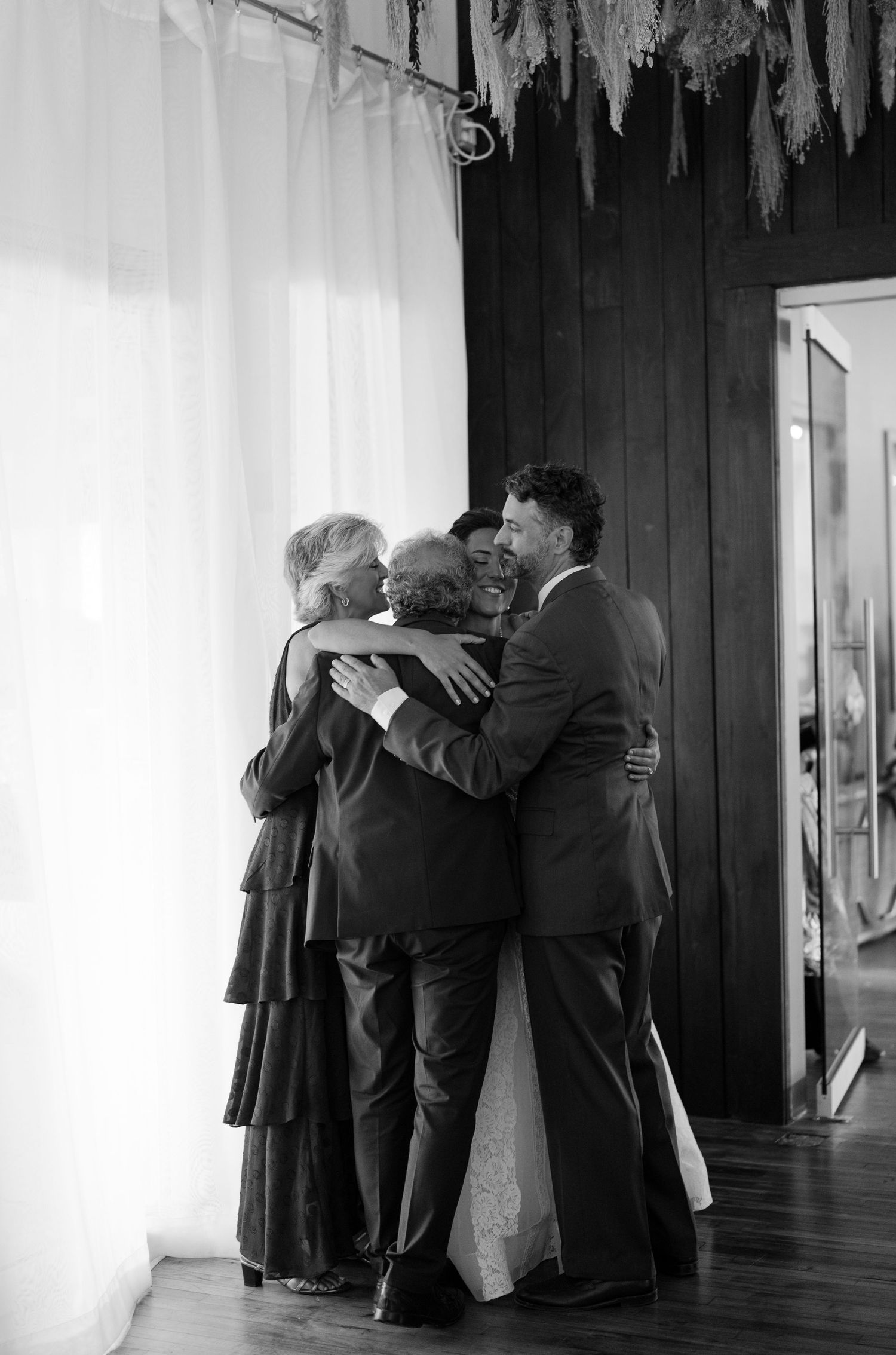 A black and white photograph of people embracing in an emotional group hug beside a curtained window.