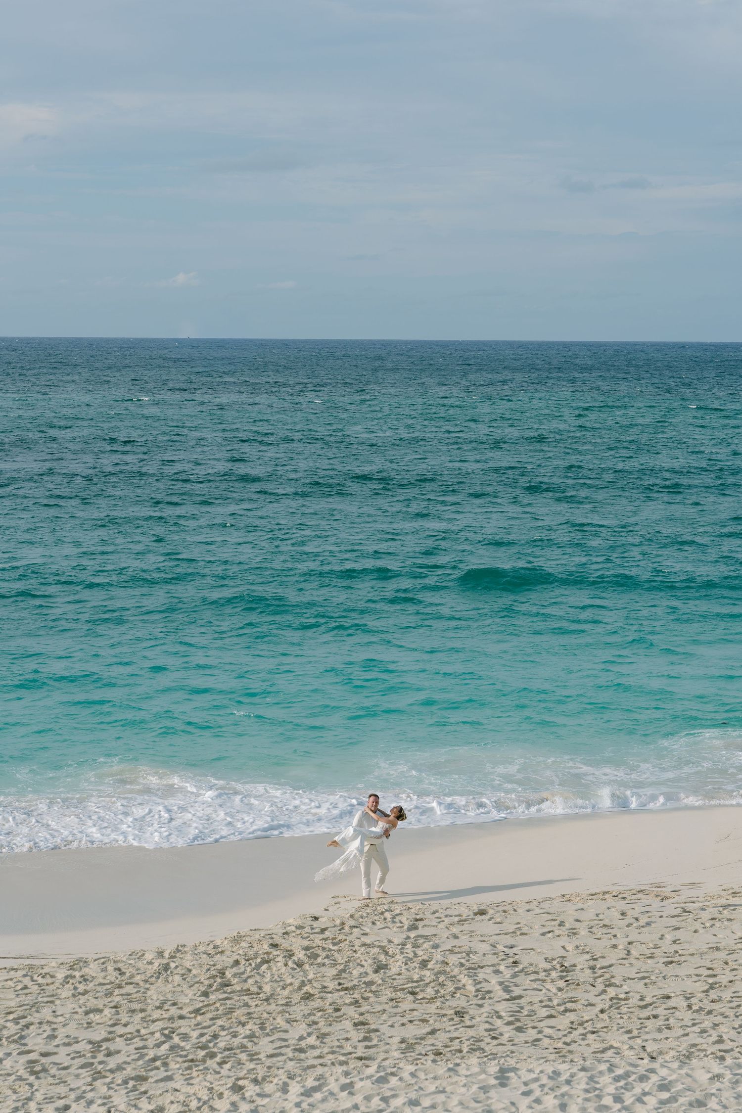 Couple in white outfits embracing on a pristine white sand beach with turquoise ocean waves in the background.